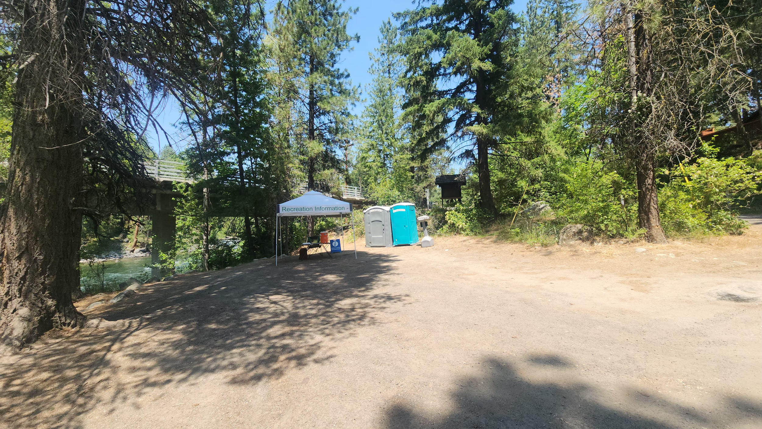 A shaded outdoor area with trees and a dirt ground, featuring a white canopy tent labeled 'Recreation Information,' a portable toilet, and a trash can.