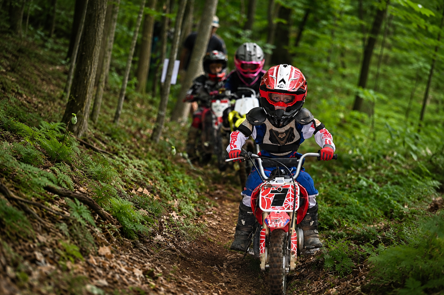 Children riding dirt bikes through a forest trail, wearing protective helmets and gear.
