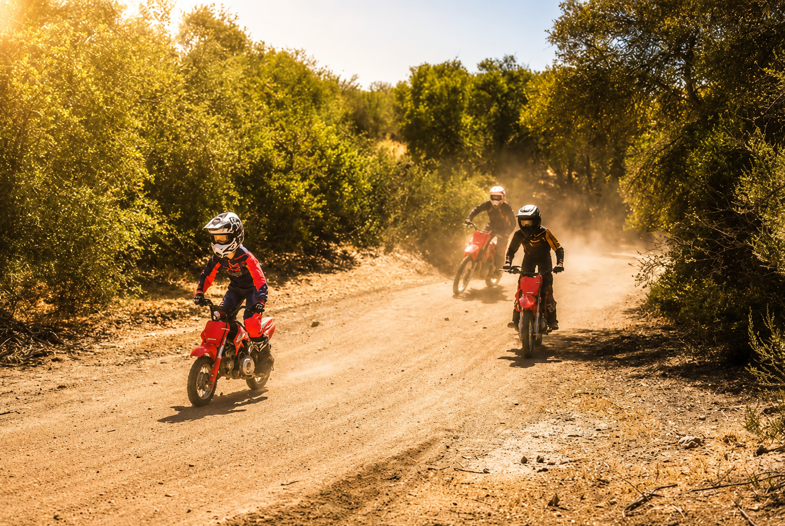 Three children wearing protective gear and helmets riding dirt bikes on a dusty trail surrounded by green bushes and trees on a sunny day.