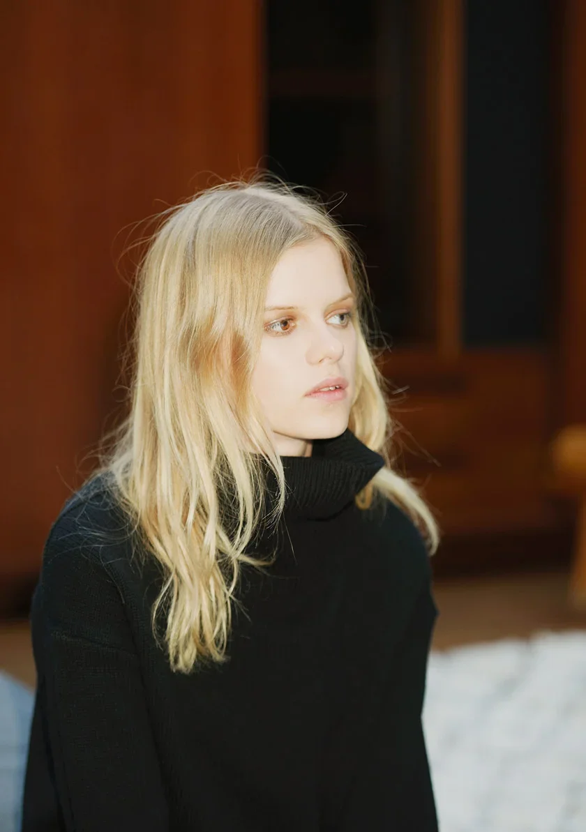 A young woman with long blonde hair, wearing a black turtleneck sweater, sitting indoors with a serious expression.
