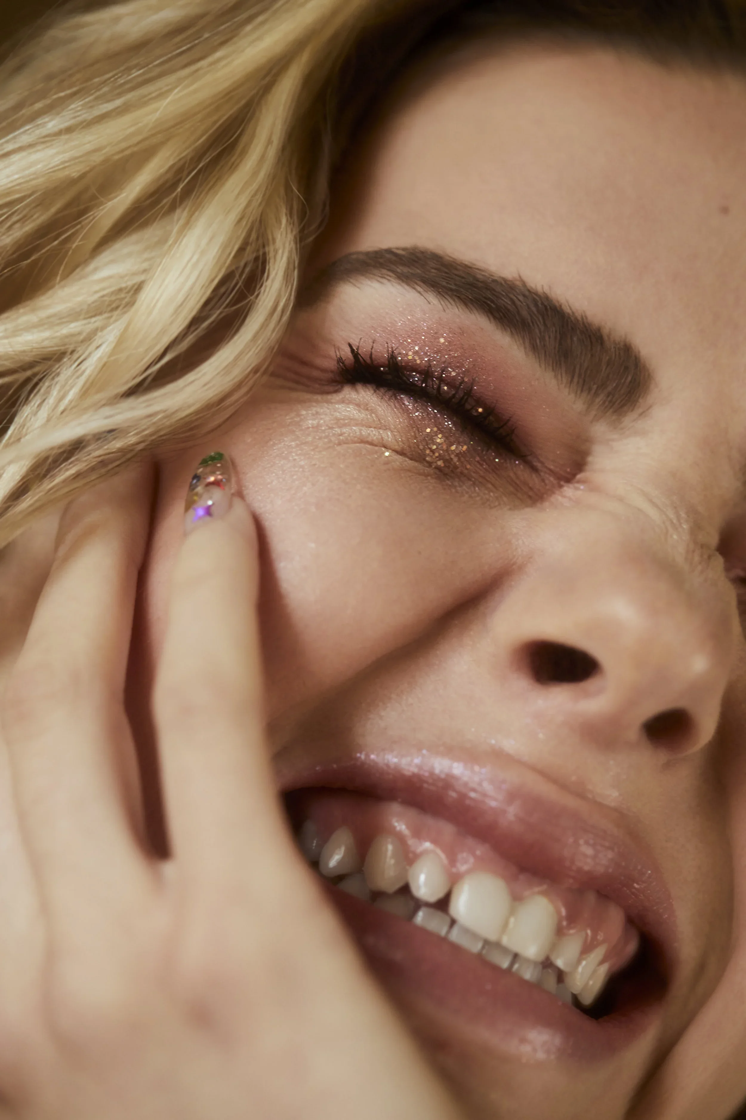 Close-up of a smiling woman with blond hair, glittery eye makeup, and white teeth, touching her cheek with her fingers.