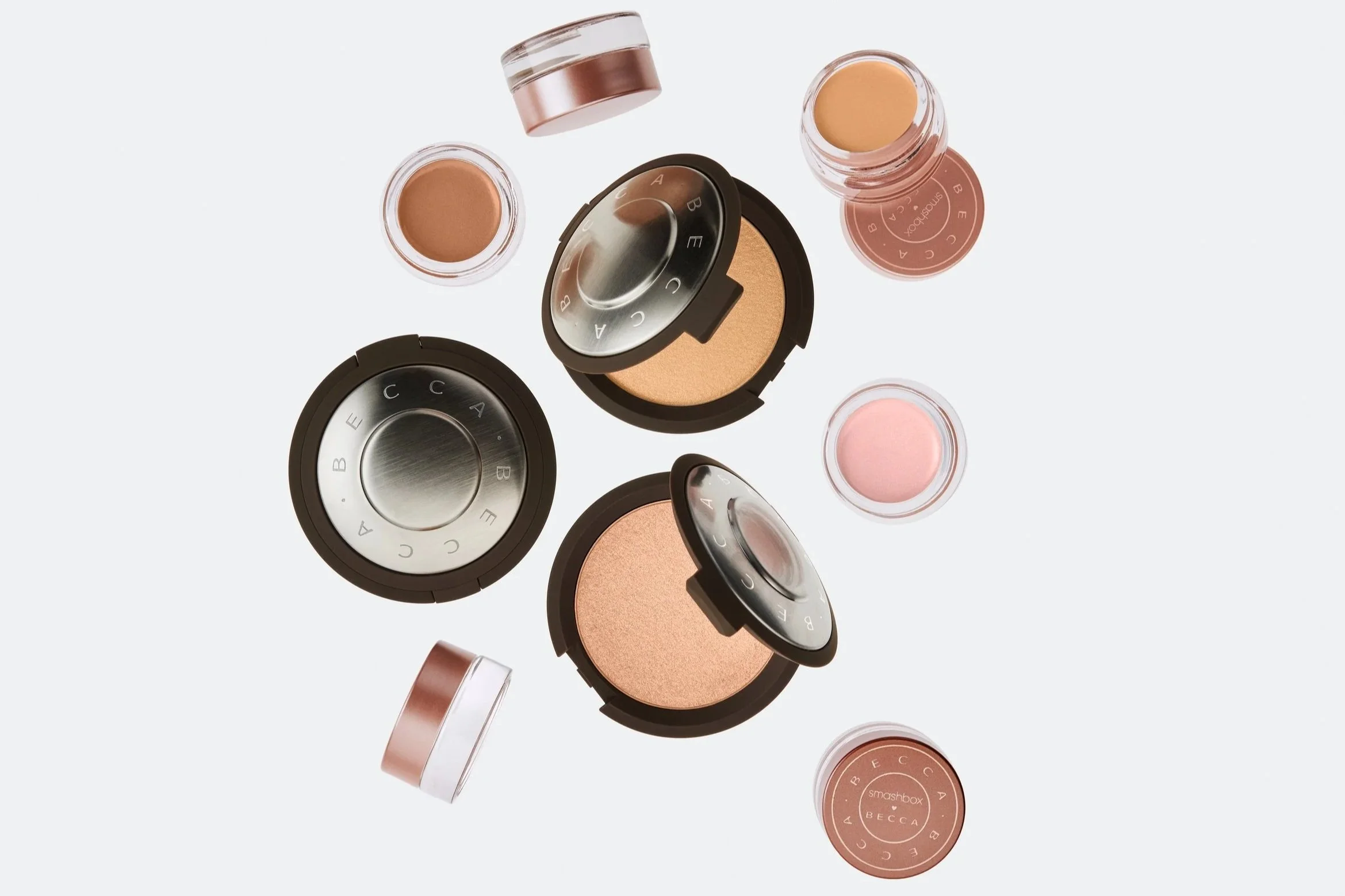 Flat lay of various makeup compact powders and jars, some open showing shades of nude, pink, and peach, with a light gray background.