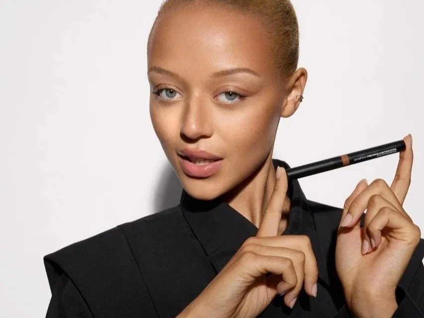 A woman with light skin and blonde hair styled in a bun, wearing a black blazer, holds a black eyeliner pencil near her face, with a playful expression and slightly raised eyebrows.