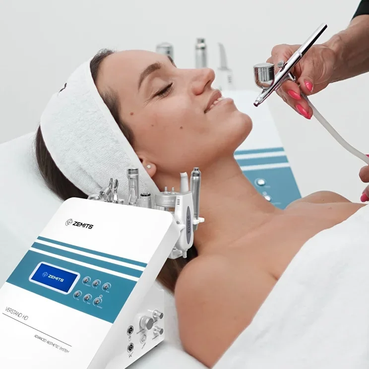 A woman receiving a facial treatment with a ZEMITS skincare device in a clinic. She has her eyes closed and a relaxed expression, with a white towel wrapped around her head.