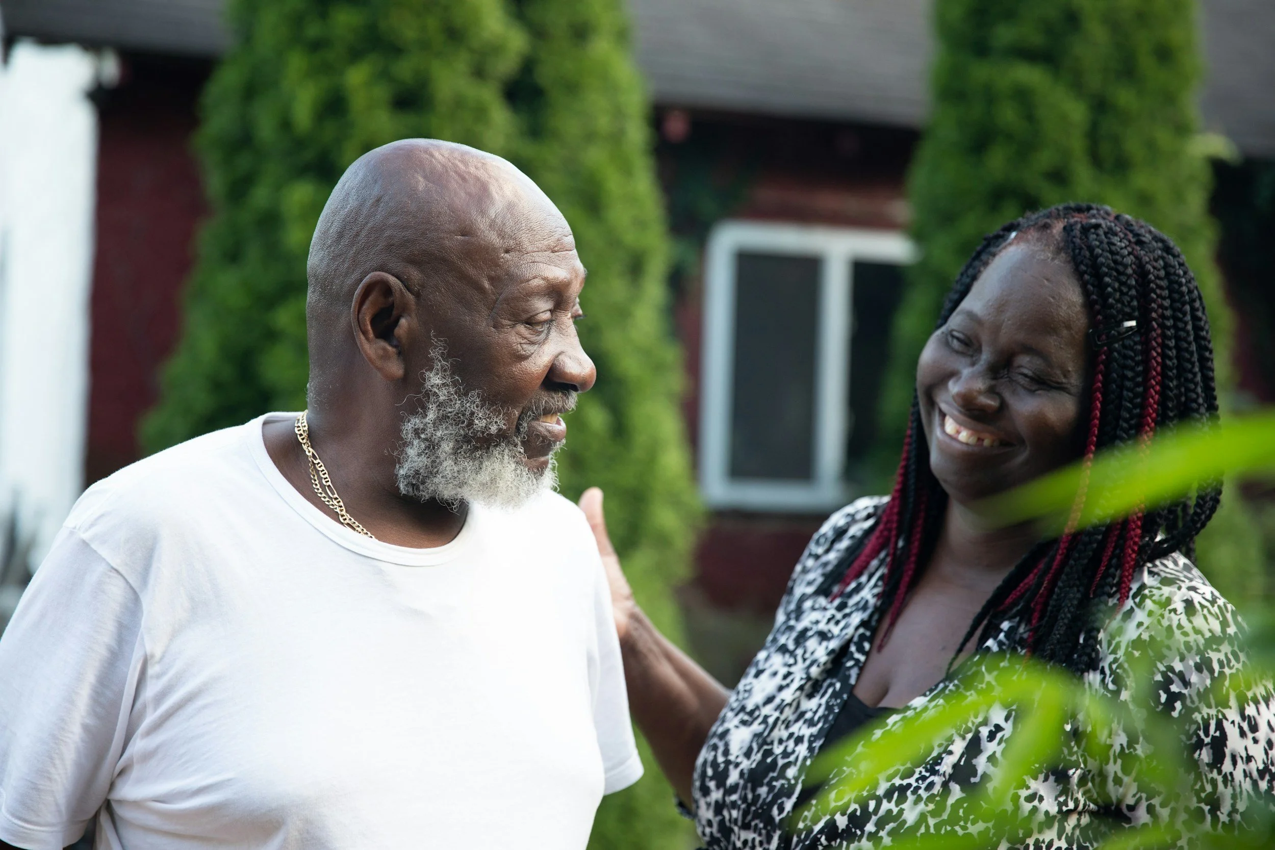 An elderly man with a gray beard and a gold chain, smiling and talking to a woman with braided hair and earrings, outdoors with green foliage in the background.