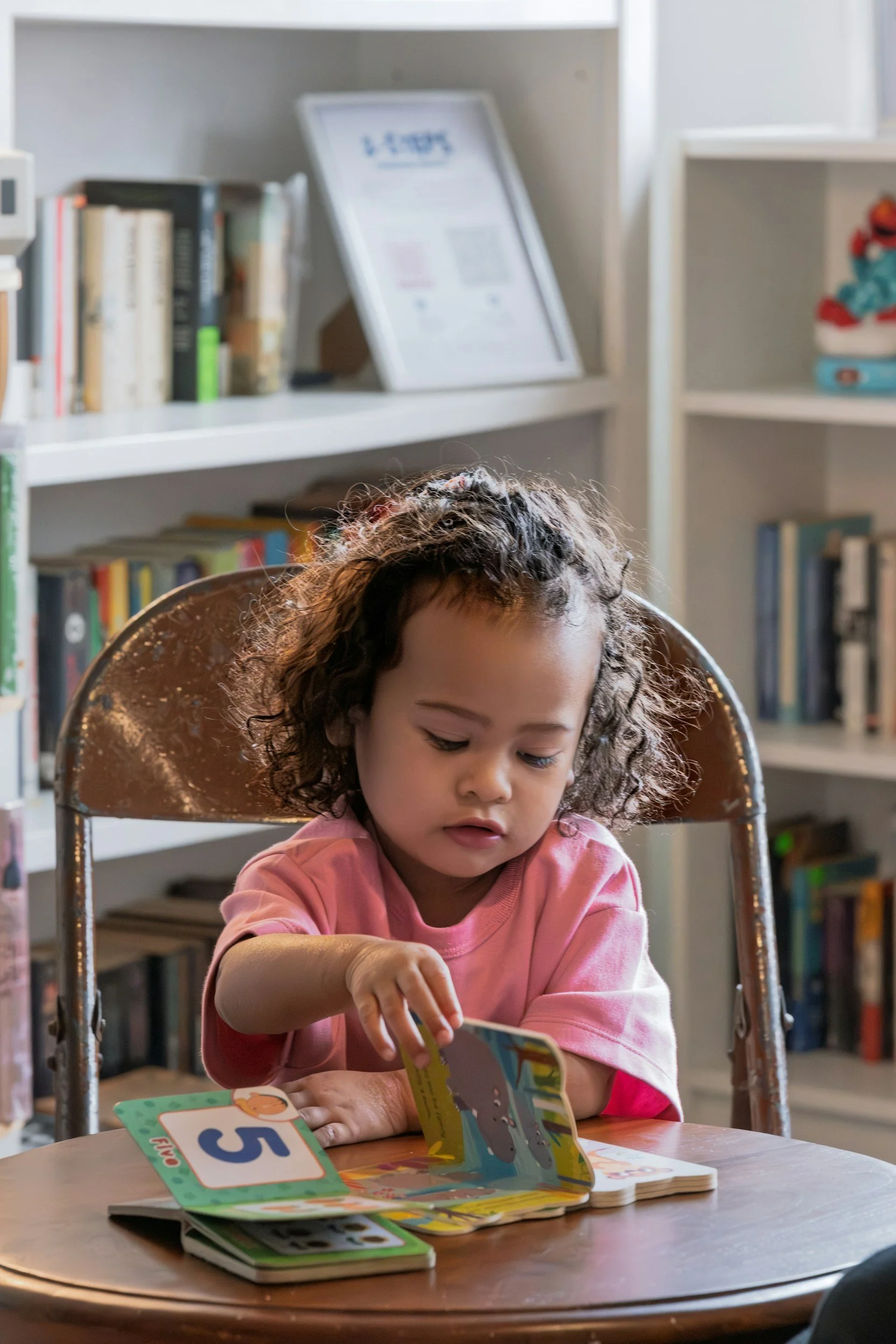A young girl with curly hair sitting at a table, reading a children's book. There are more books and a numbered card on the table. The background includes bookshelves and a framed picture.