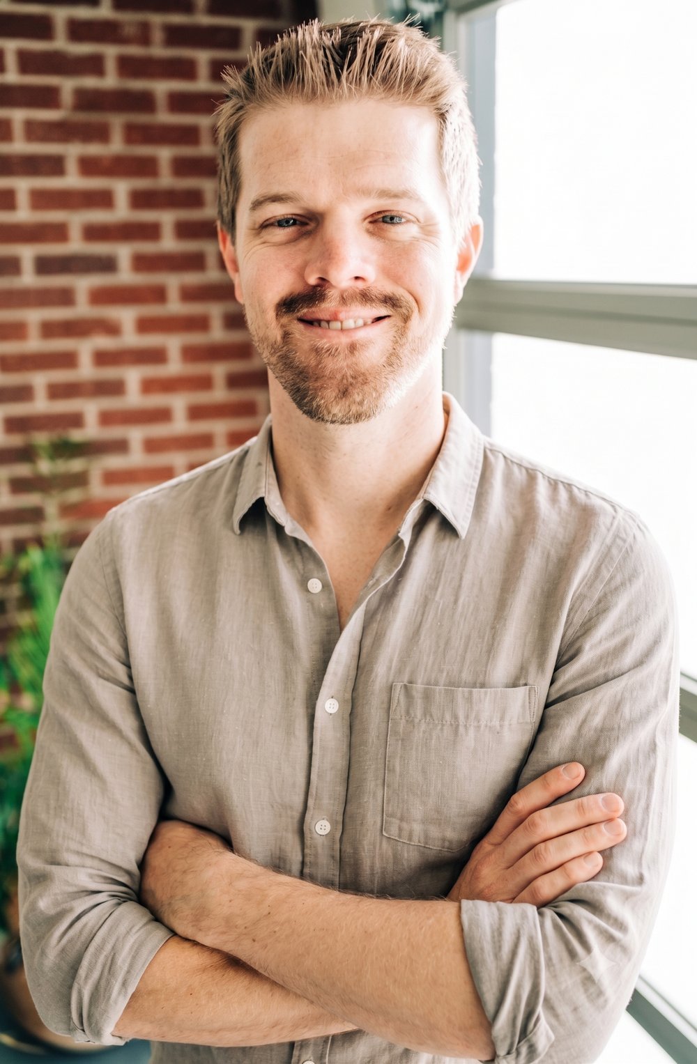 Brian Geier , A smiling man with light brown hair and a beard standing with arms crossed near a window with a brick wall background.