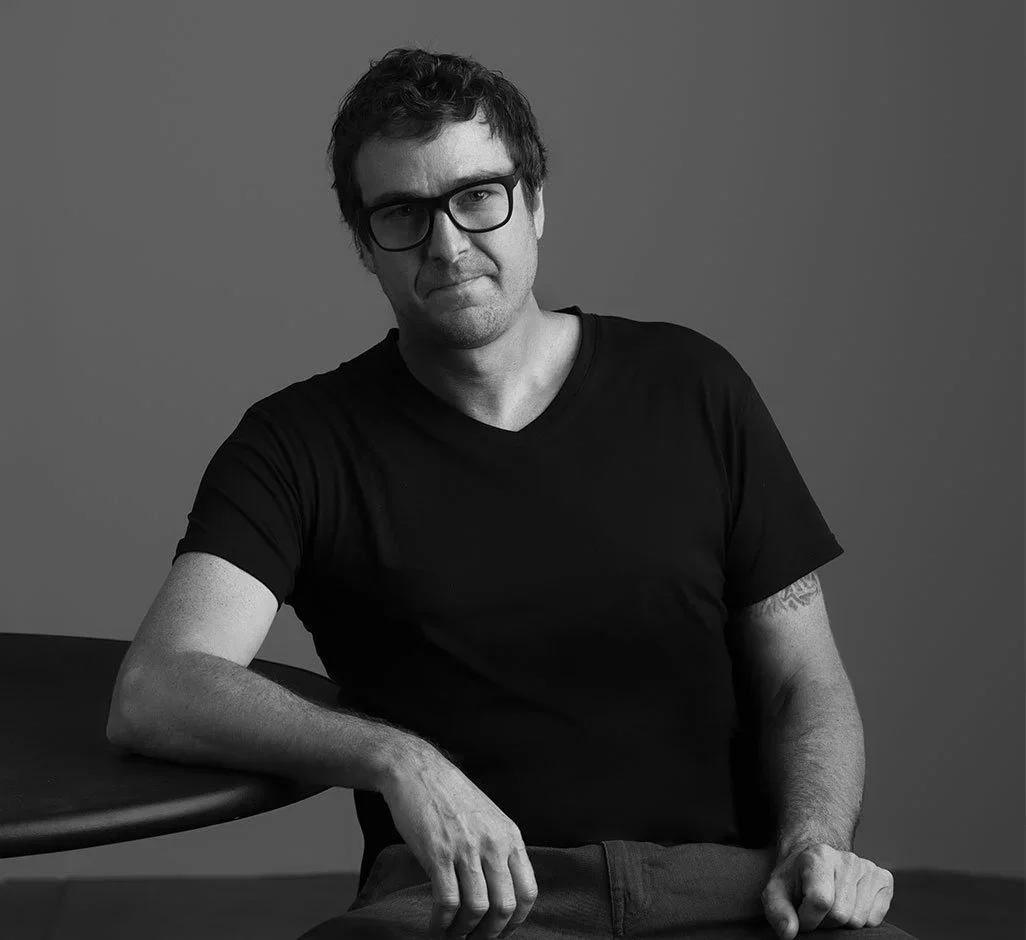 Andy Morter, a man with glasses and dark hair, wearing a black T-shirt, sitting at a desk with his arm resting on it, in a black-and-white portrait against a plain background.