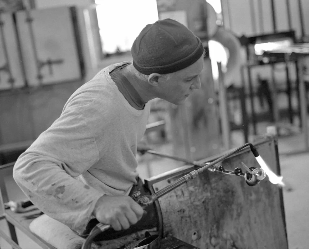 John Chiles, a glassblower wearing a beanie using a blowtorch to shape glasswork in a workshop.