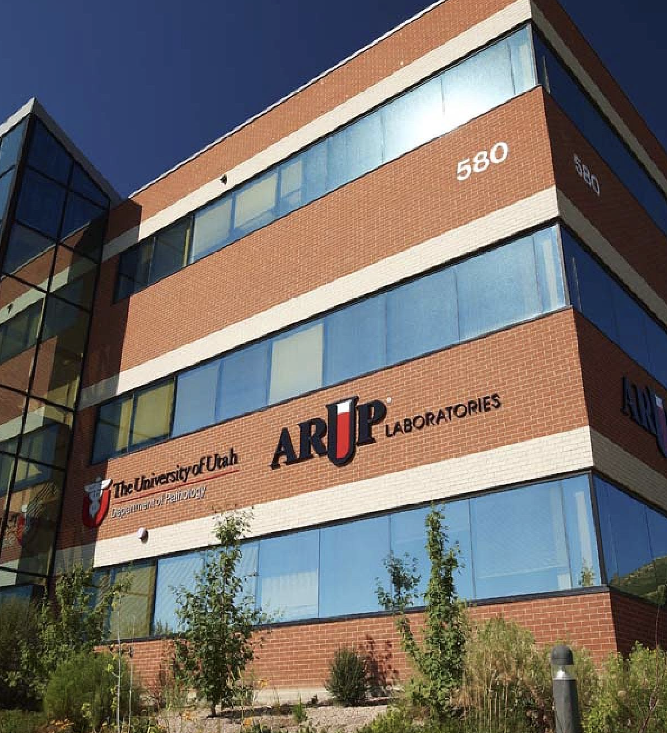 Exterior view of a modern brick building with large glass windows, displaying signs for The University of Utah, ARUP Laboratories, and the Department of Pathology. The building number 580 is visible on the corner, and there is landscaping with small trees and shrubs in front.