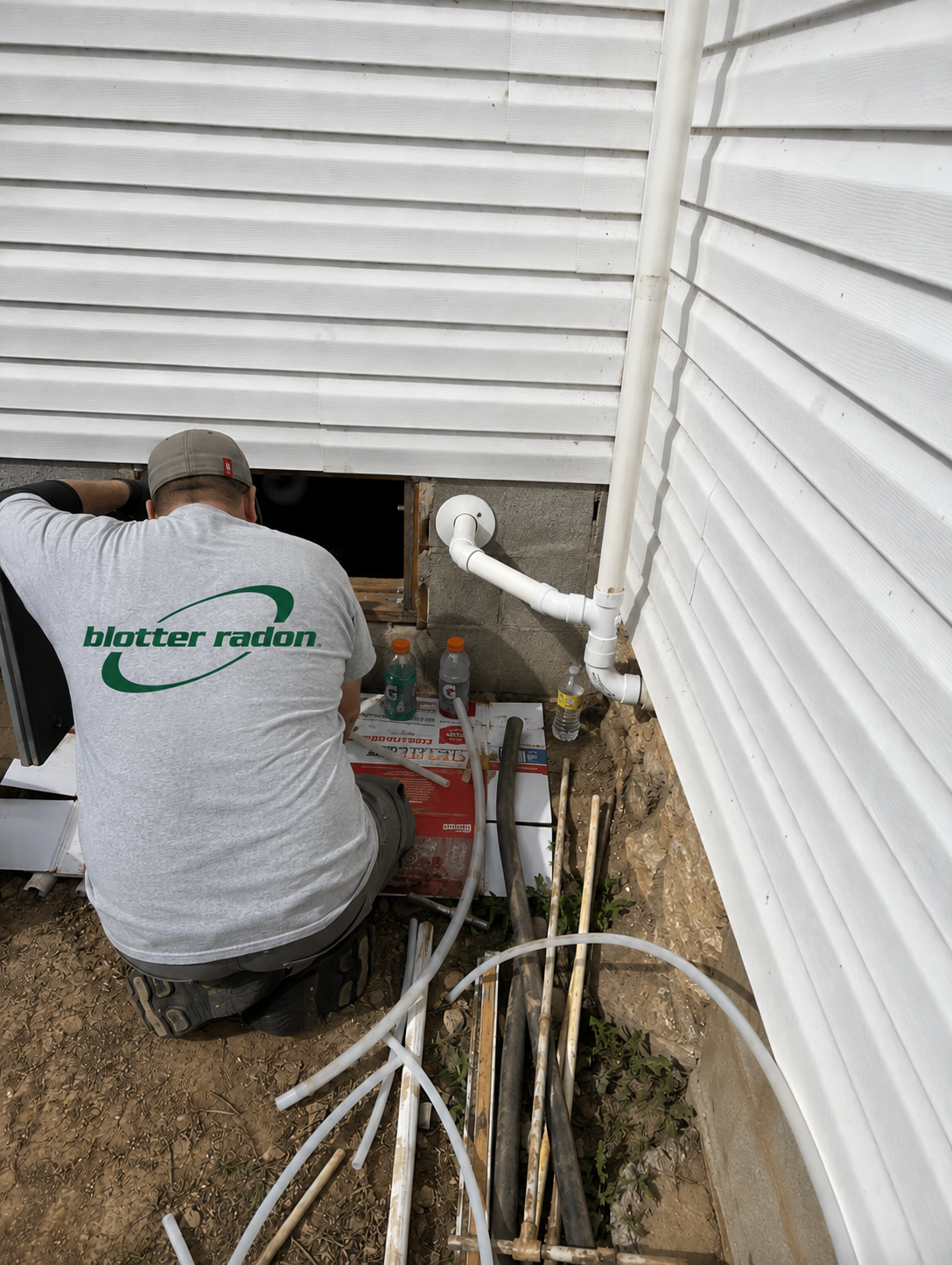 A worker installing or repairing plumbing pipes outside a house with white siding. The worker is kneeling and has a gray cap and a shirt with 'blotter radon' written on the back. There are various pipes, bottles, and tools on the ground.