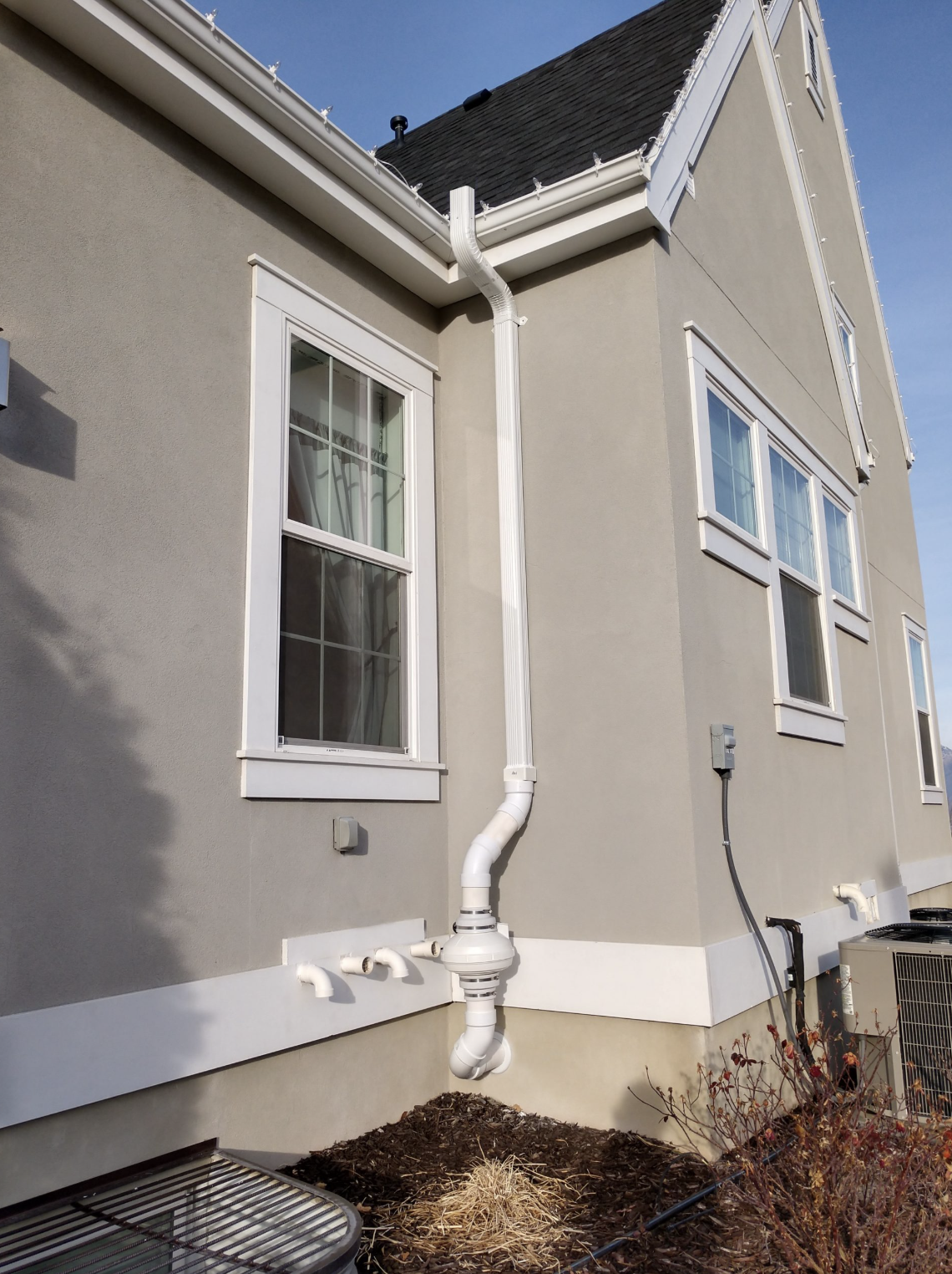 Side of a house with multiple white-framed windows, beige exterior walls, and visible piping for drainage and utility connections, set against a clear blue sky.