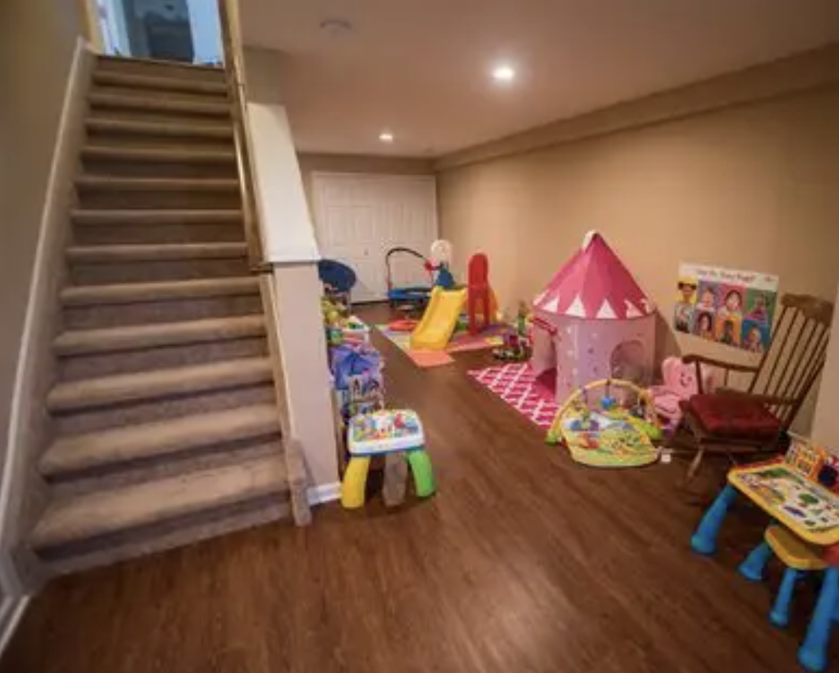 A playroom with a staircase on the left, and various children's toys, a pink tipi tent, play mats, and a wooden rocking chair on the right side.