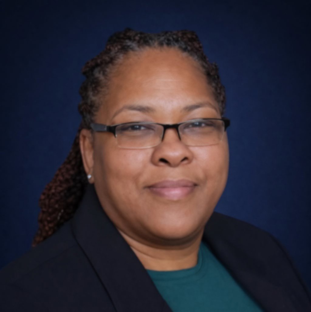 A woman with glasses and braided hair, wearing a dark blazer and a teal shirt, smiling at the camera against a dark blue background.