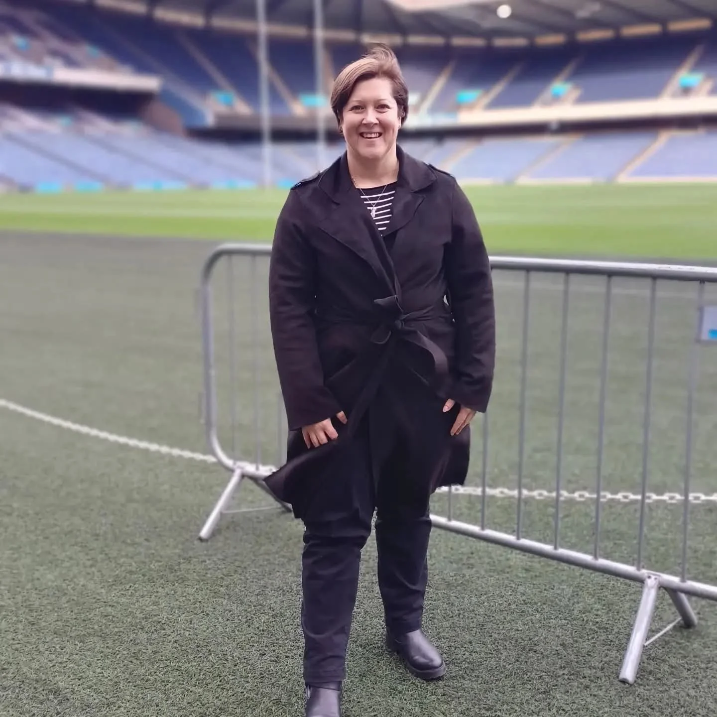 A woman standing on a sports field with stadium seating in the background, wearing a black coat, black pants, and black boots, smiling at the camera.