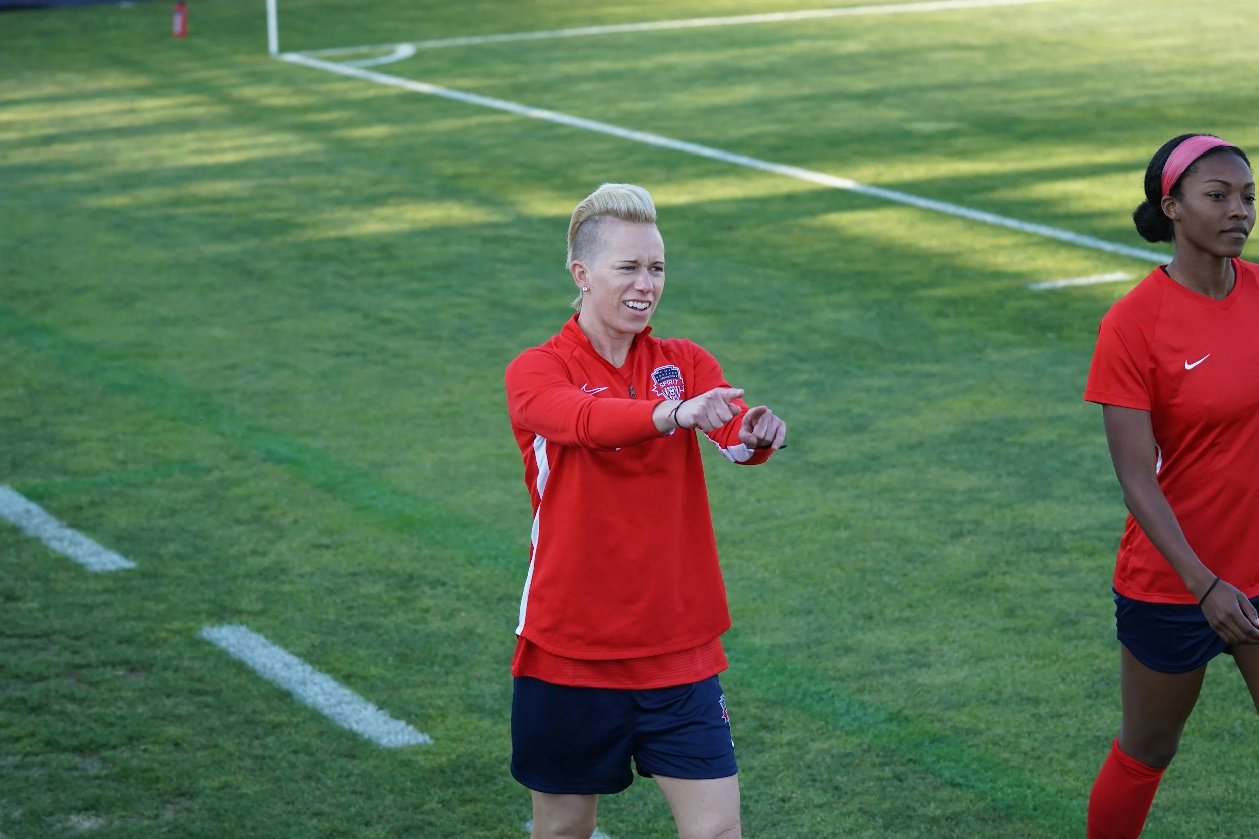 A woman with a platinum blonde undercut hairstyle wearing a red sports jacket with an American flag emblem is pointing with her right hand on a soccer field. She appears focused and is surrounded by other players, with the grassy field and white sideline markings visible in the background.