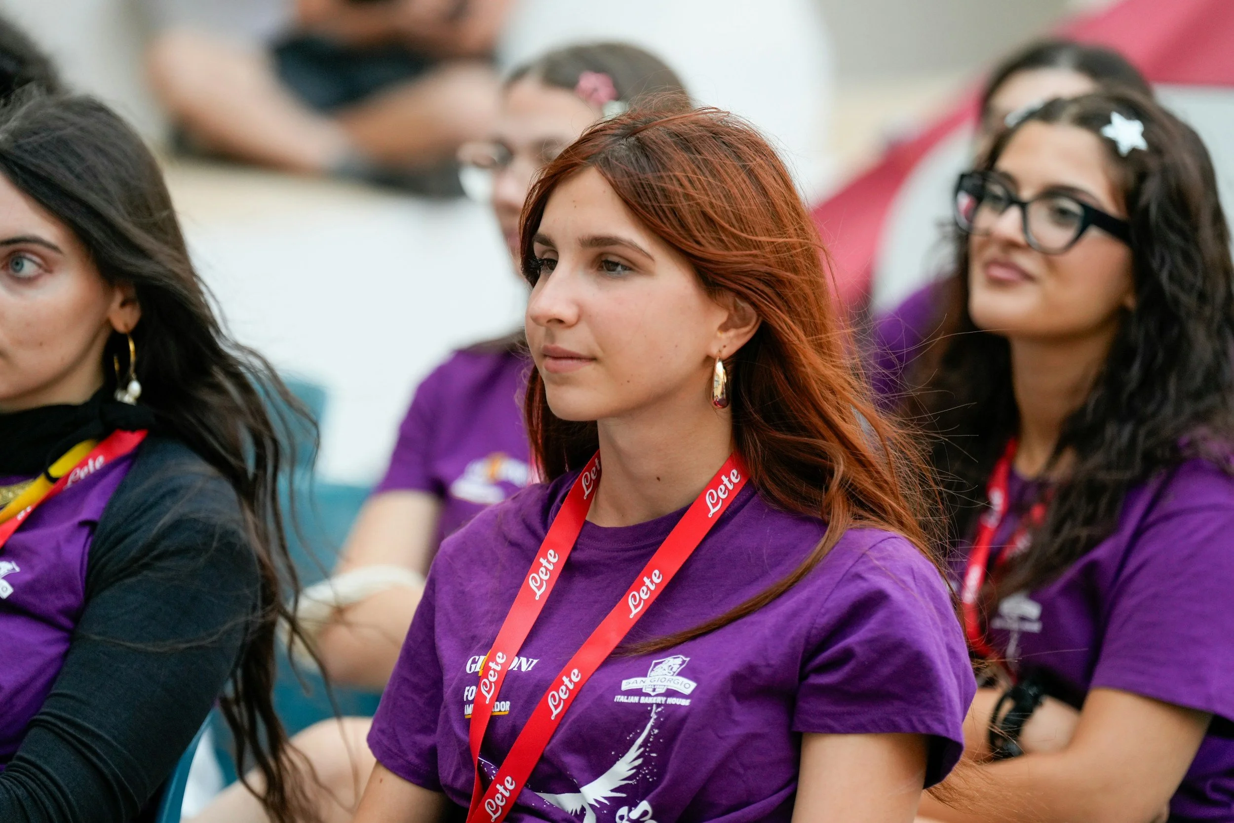 A group of young women sitting together, wearing purple T-shirts with logos, and red lanyards around their necks. They appear to be attending an event or gathering indoors.