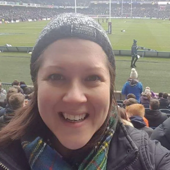 Woman at a sports stadium, smiling at the camera, wearing a Glasgow Warriors supporter hat, with the field and spectators in the background.