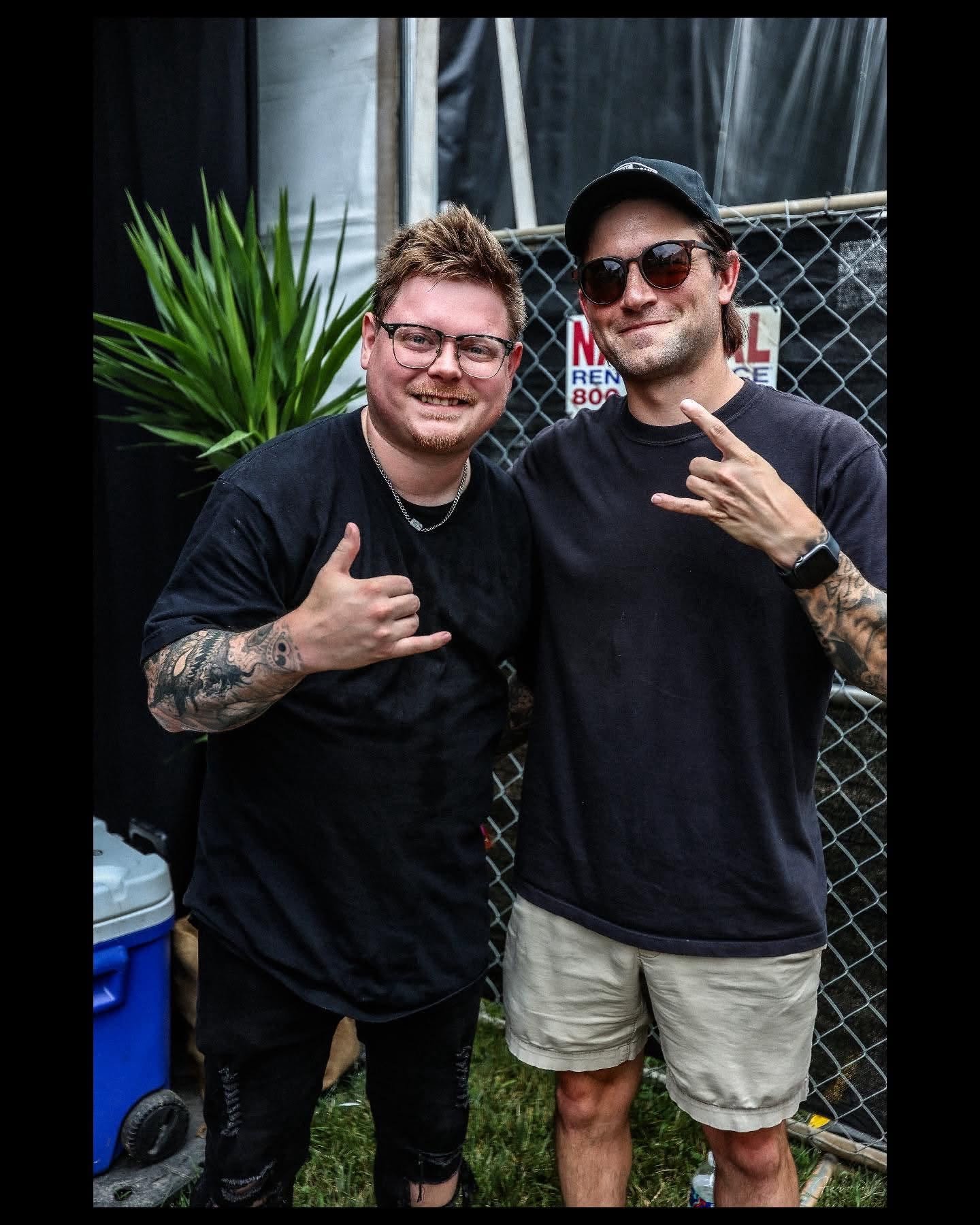 Two men standing outdoors near a chain-link fence, with one wearing glasses, a black shirt, and tattoos, giving a thumbs up, and the other wearing sunglasses, a black cap, a black shirt, and beige shorts, making a hand gesture. There is a green plant behind them and a sign on the fence.