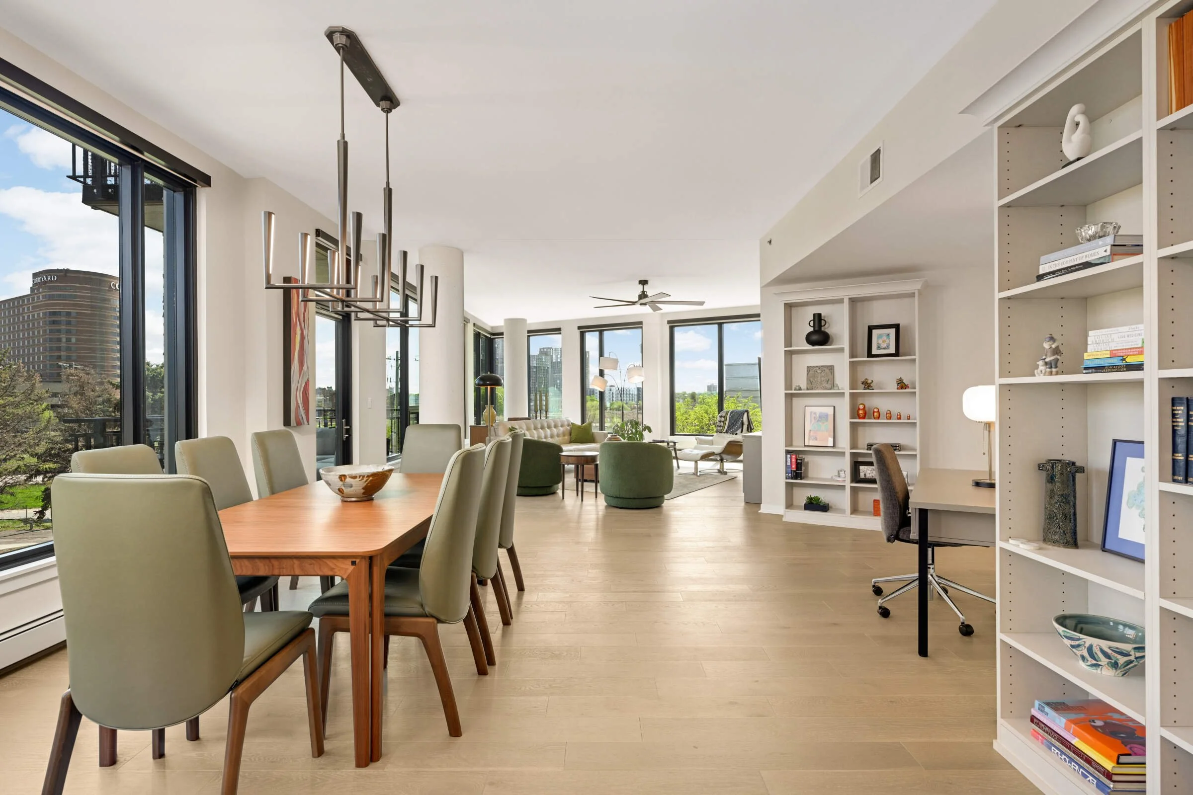 Open-concept living and dining area with large windows, modern chandelier, multiple seating options, bookshelf, and decor.
