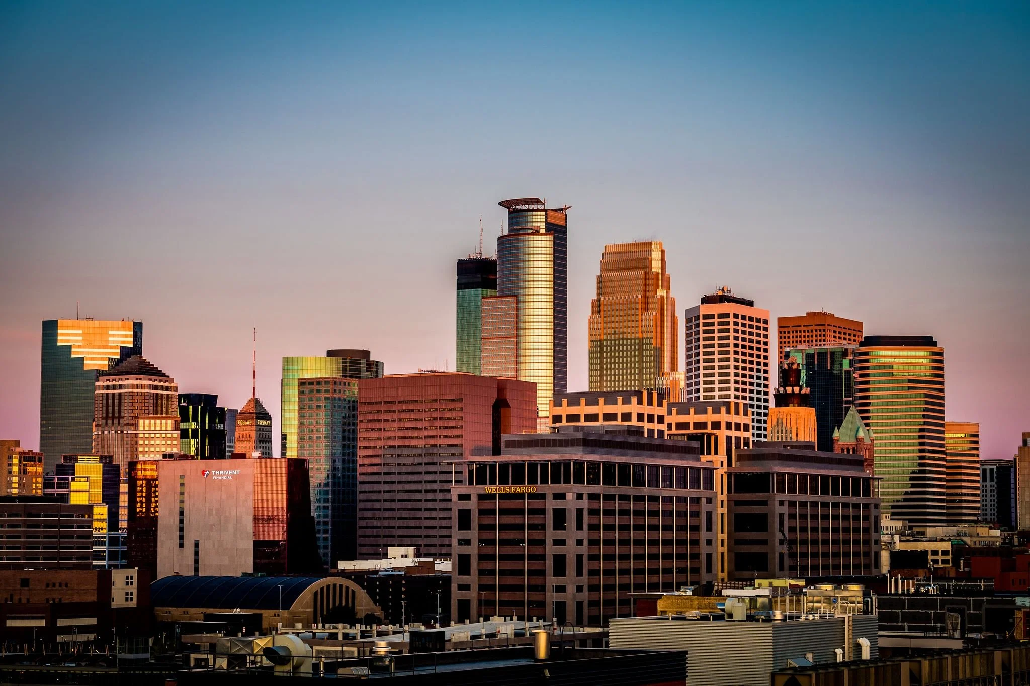 Minneapolis, Minnesota skyline from Mill District with tall modern skyscrapers during sunset, featuring glass buildings reflecting sunlight, with a clear sky overhead.