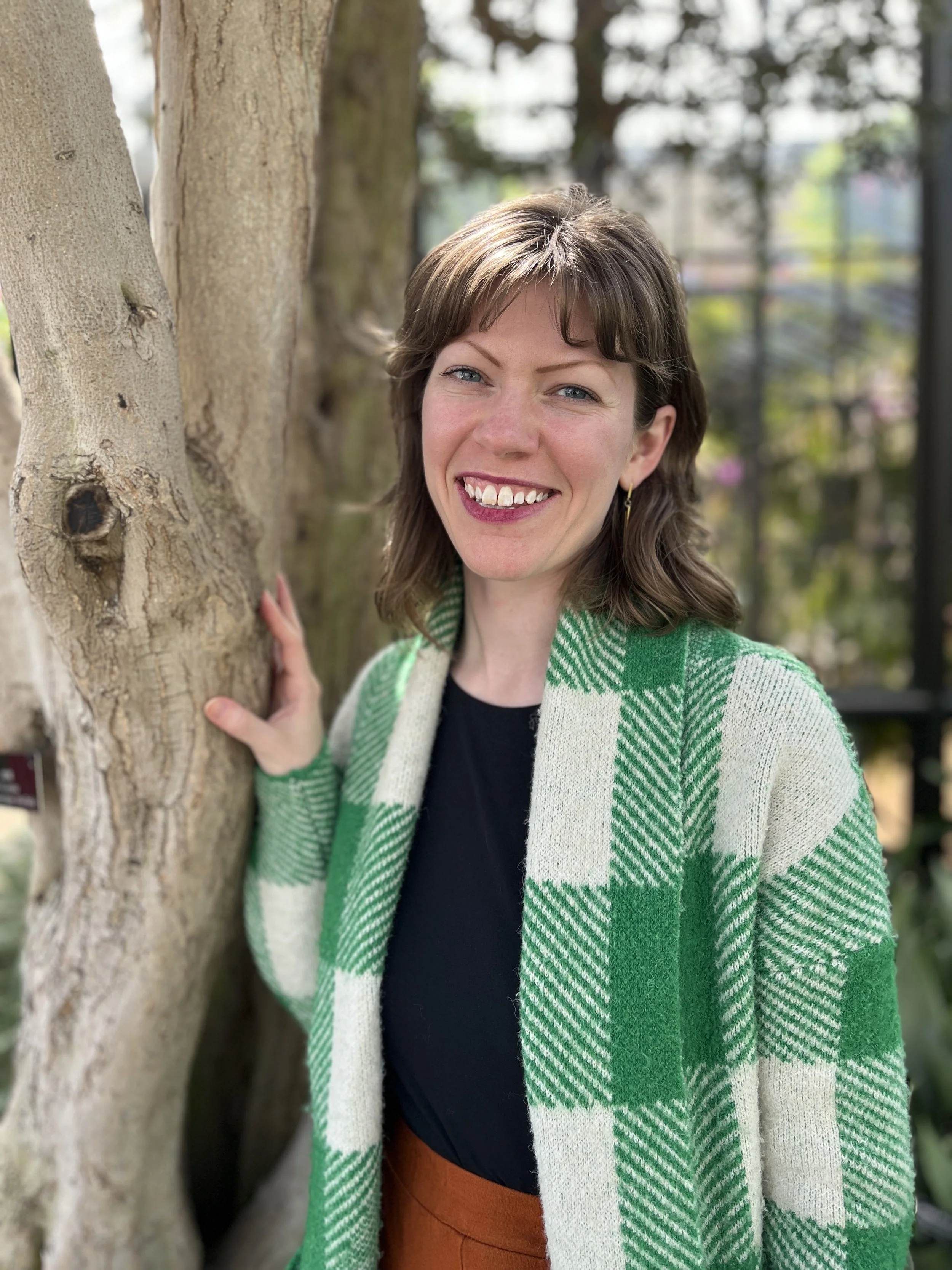 A woman with brown hair and blue eyes smiling, wearing a green and white plaid cardigan over a black top, standing outdoors beside a tree with textured bark, with blurred trees and architectural elements in the background.