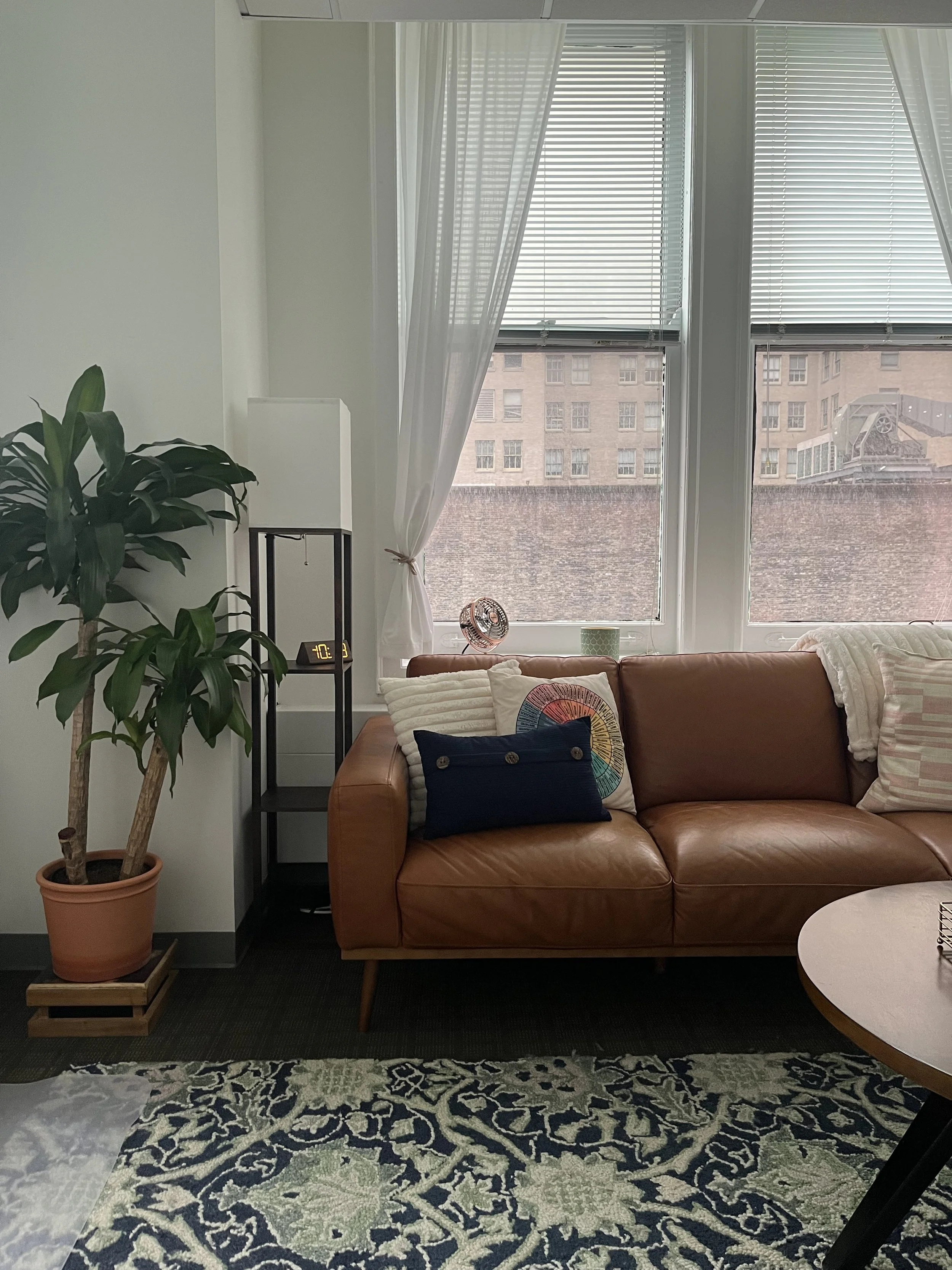 Interior of a living room with a tan leather sofa, decorative pillows, a small round white table, a tall potted plant on a wooden stand, a side shelf with an alarm clock, and large windows with blinds and sheer curtains looking out onto a brick building.