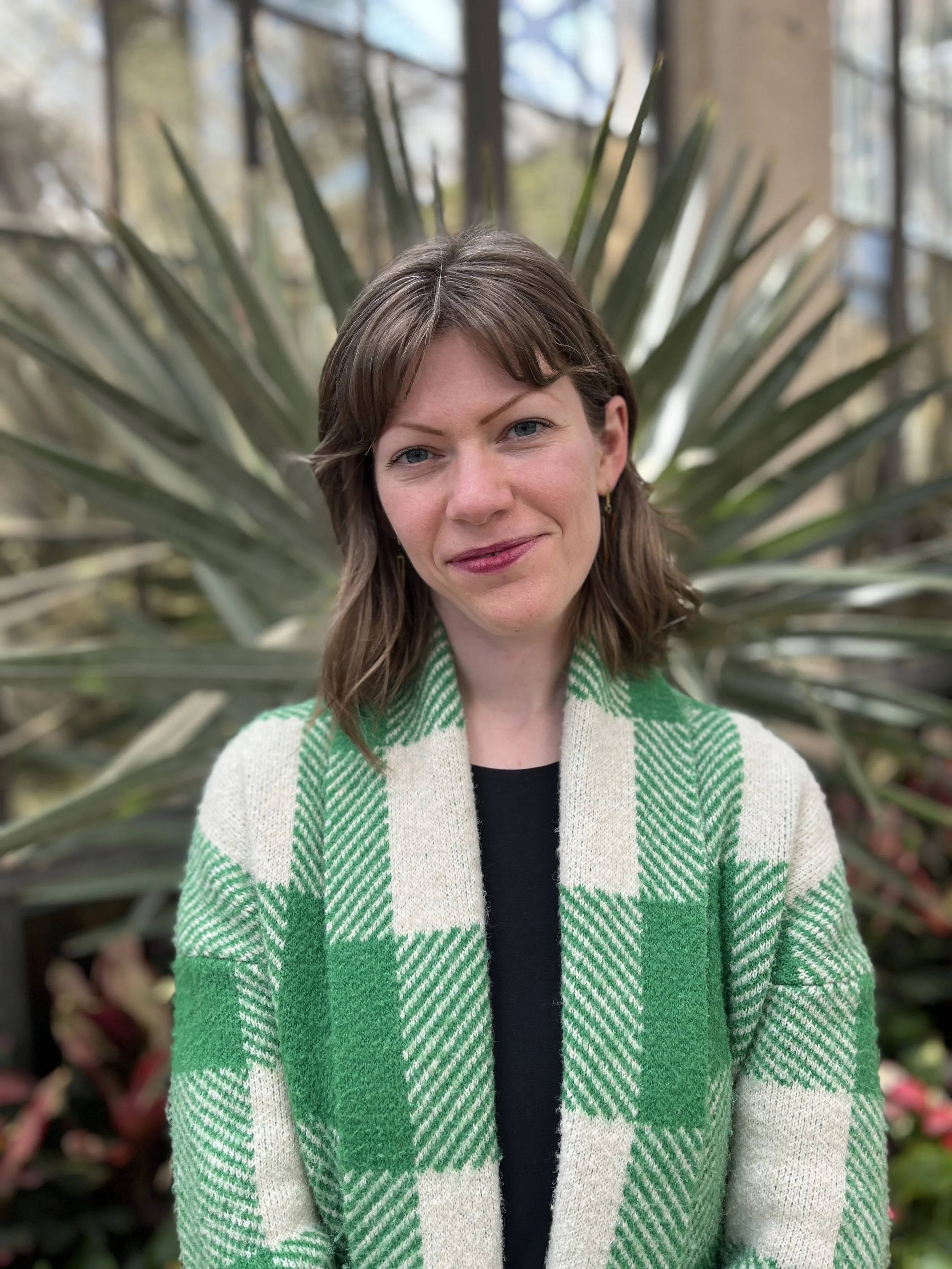 Portrait of a woman with brown hair and blue eyes, wearing a green and white striped sweater, standing outdoors with a large plant and buildings in the background.