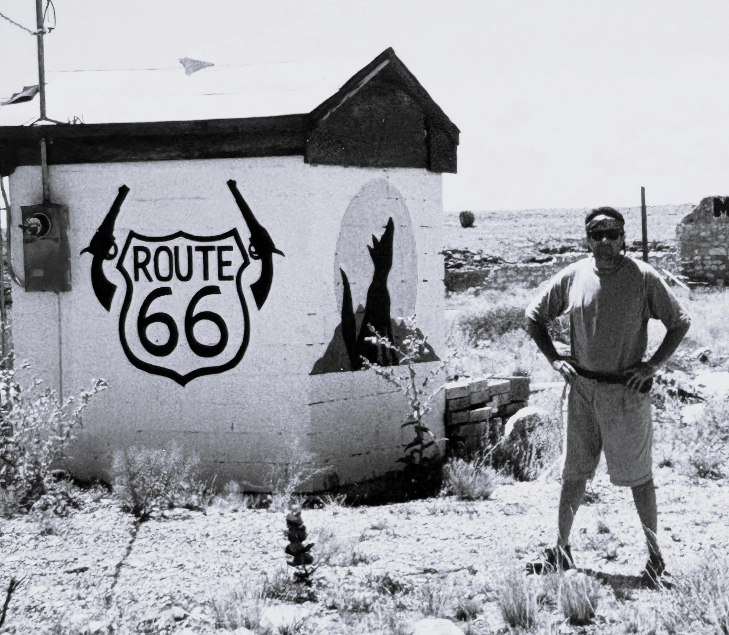 A snowy landscape with a small building or shed, a man wearing a hat and glasses, and a Route 66 sign with two guns crossed behind it.
