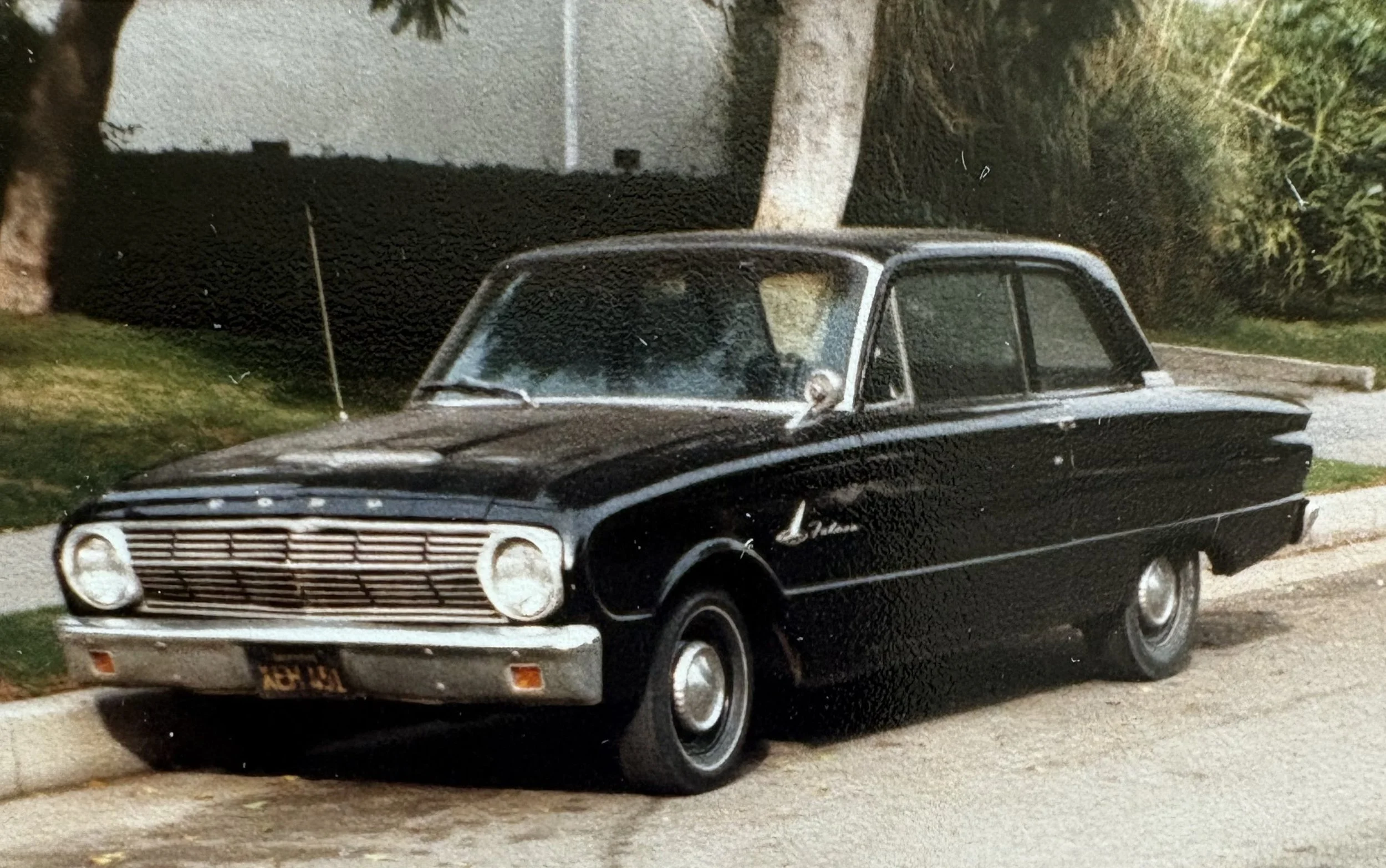A vintage black two-door car parked on a suburban street with a tree and house in the background.