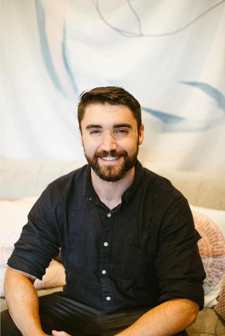 A man with dark hair and a beard, wearing a black button-up shirt, sitting on a couch with pillows, smiling at the camera.