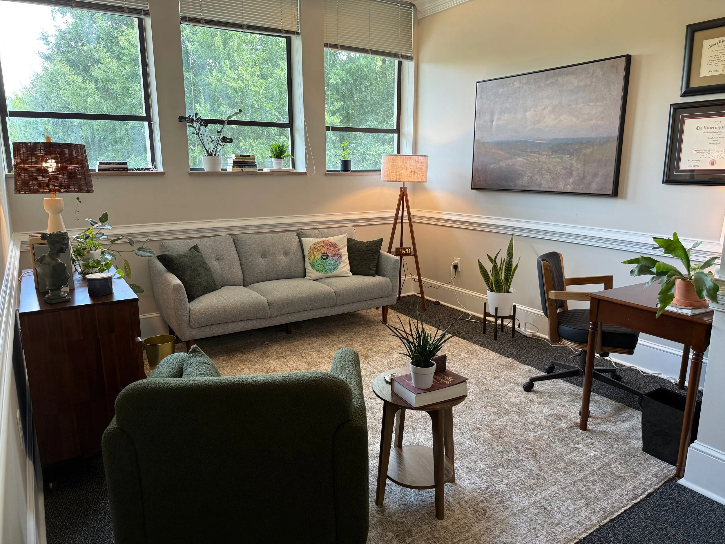 A cozy living room with large windows showing green trees outside. The room has a light gray sofa with dark cushions, a wooden side table with plants and decor, a standing lamp, a carpeted floor, and framed artwork on the wall. There is also a small round table with a potted plant and some books.
