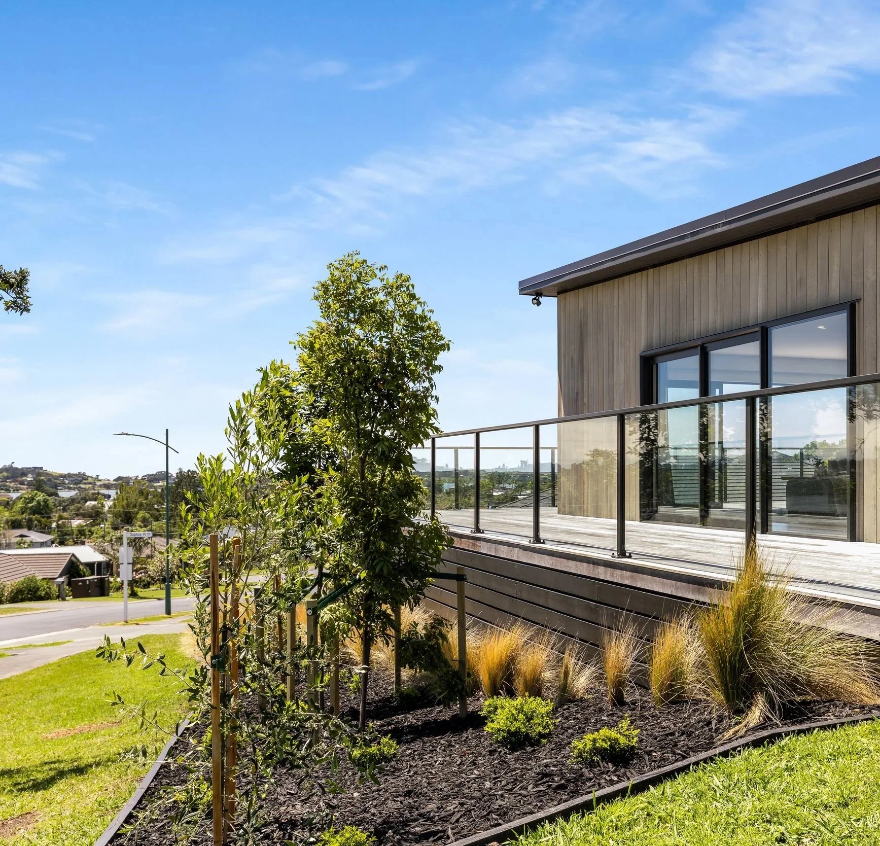 Modern house with cedar exterior walls and large glass windows, surrounded by a landscaped yard with trees, tall grasses, and a glass railing on a bright, sunny day.