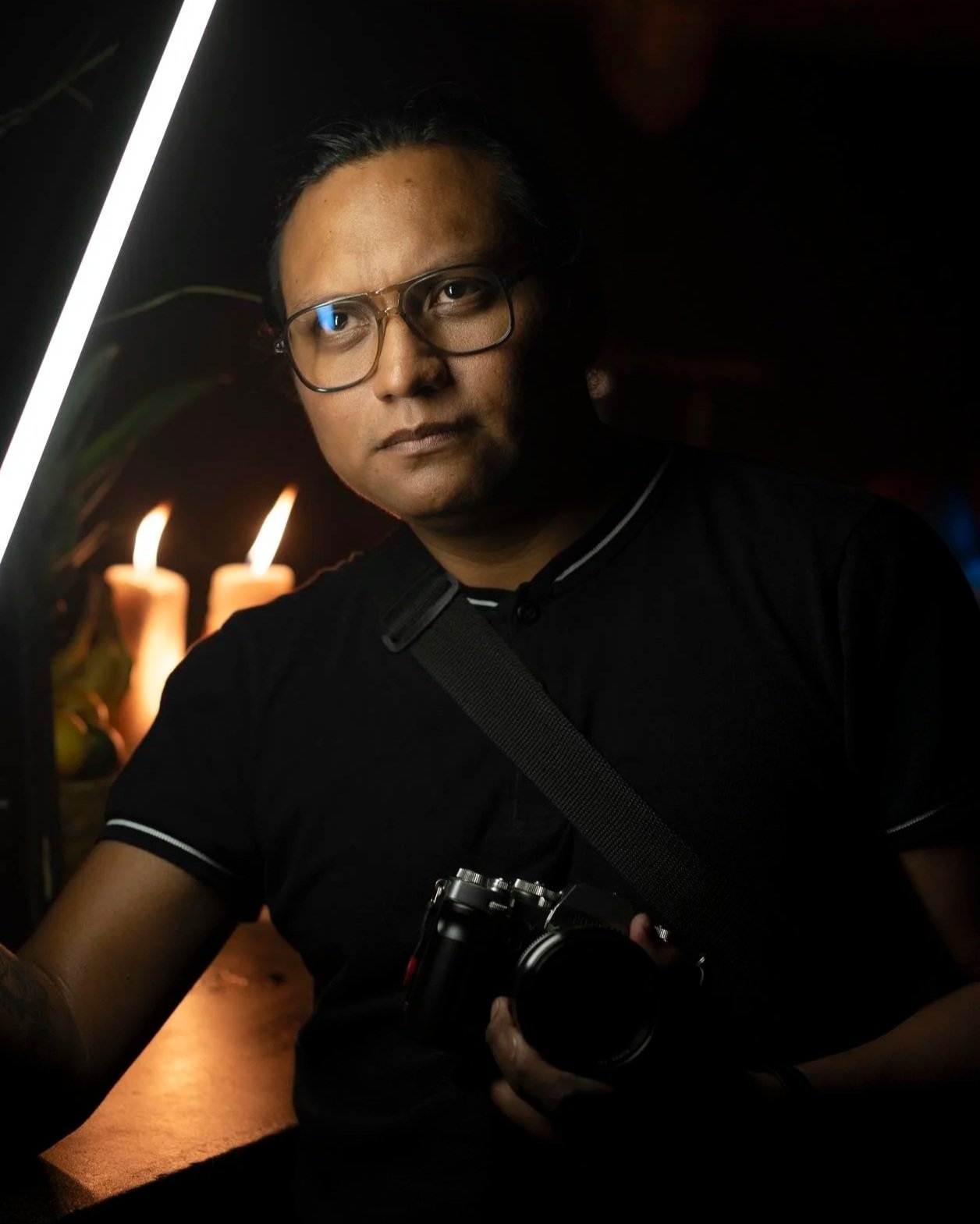 Man wearing glasses holding a camera in a dimly lit setting with candles in the background.