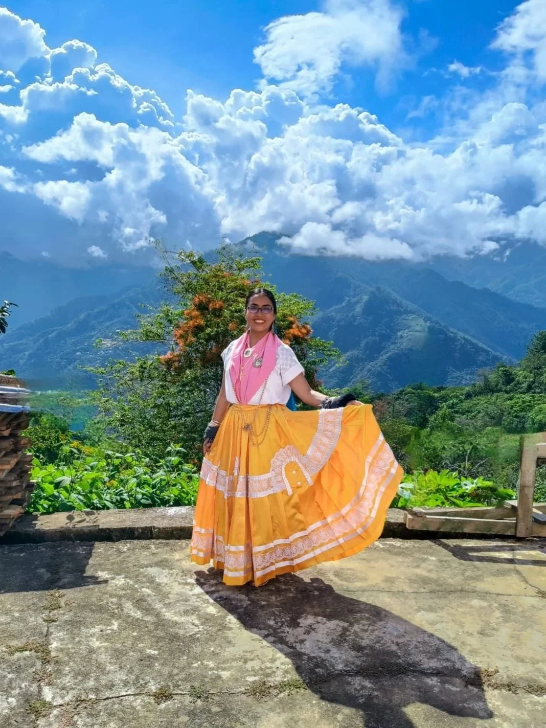 A China Oaxaqueña in traditional clothing standing outdoors against a mountainous landscape with clouds in the sky.