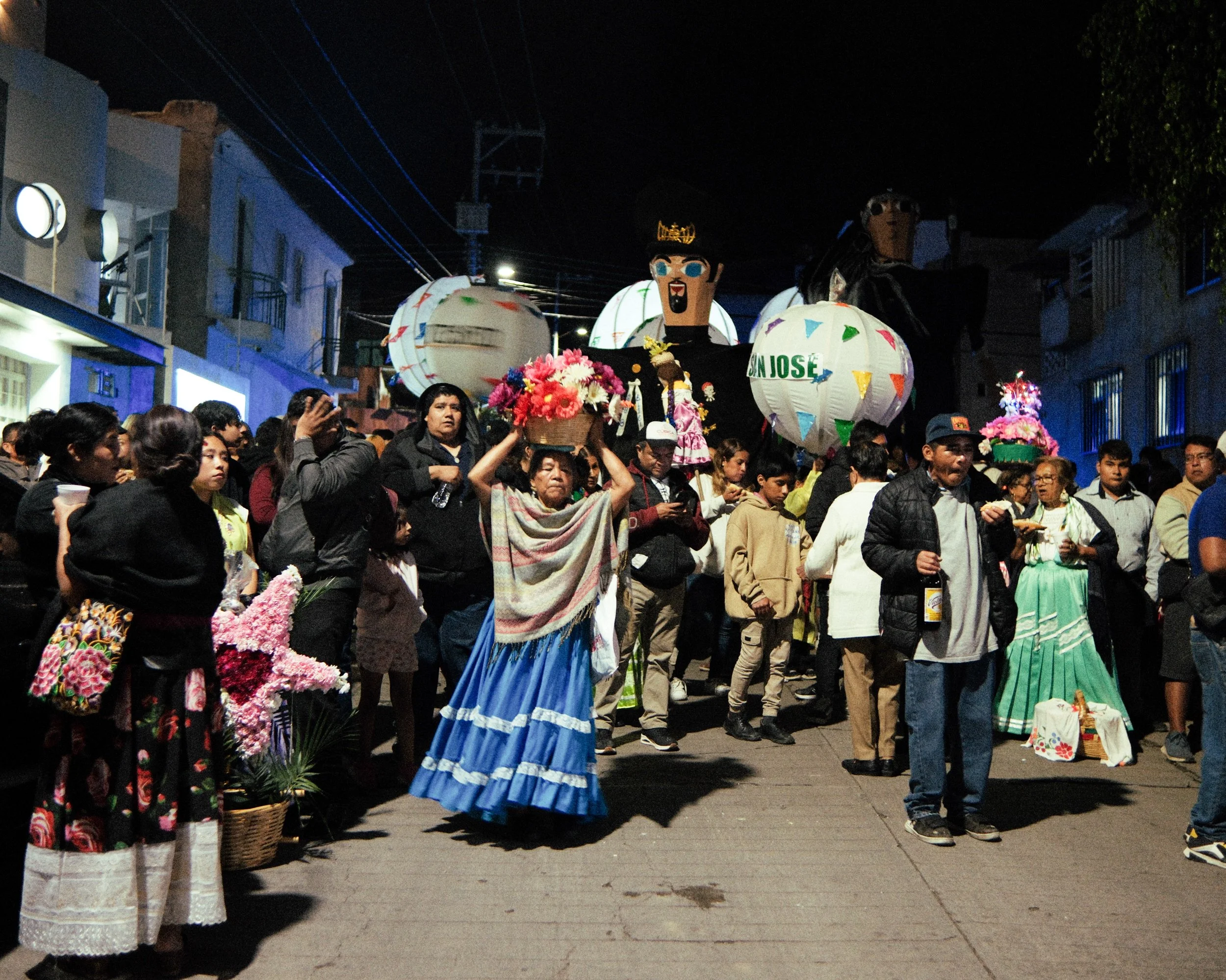 Nighttime street scene with a parade featuring large marmotas, surrounded by crowd of people, some in traditional clothing, with the cuty in the background.