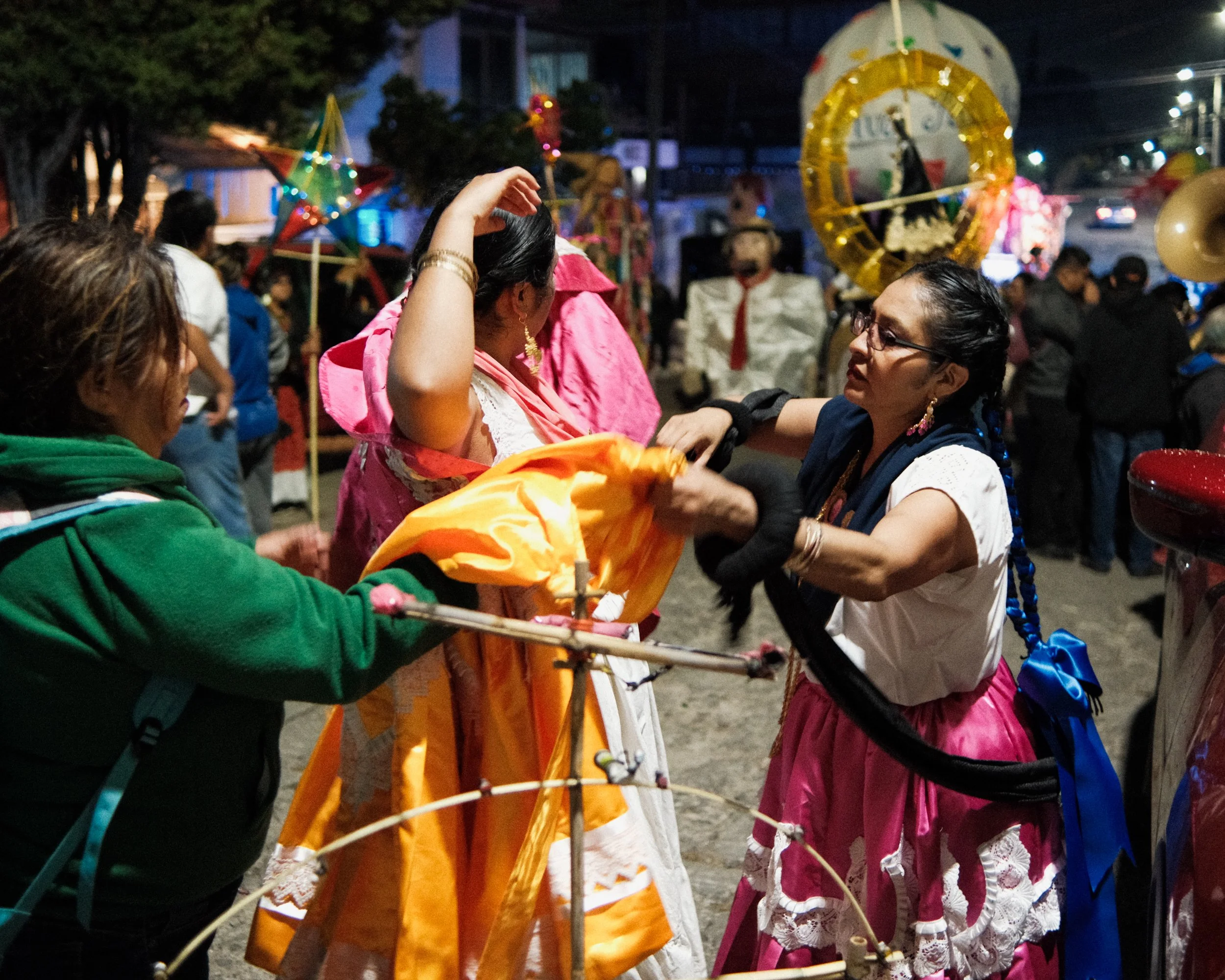 People preparing for a traditional celebration or calenda at night, with some wearing colorful skirts, accessories, and traditional clothing, dressing as china oaxaqueñas with marmotas and festive objects in the background.