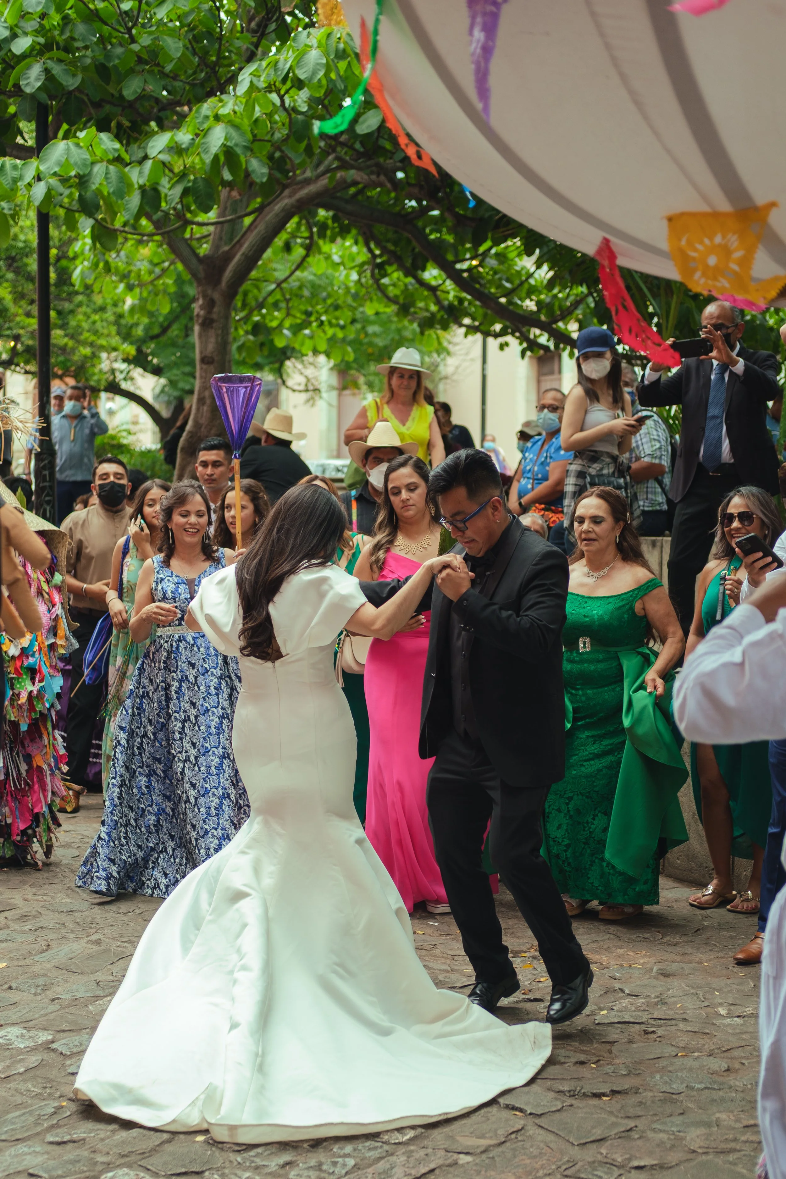 People at an outdoor calenda, with a bride dancing with a groom in a black suit, surrounded by guests, marmotas and flower decorations.