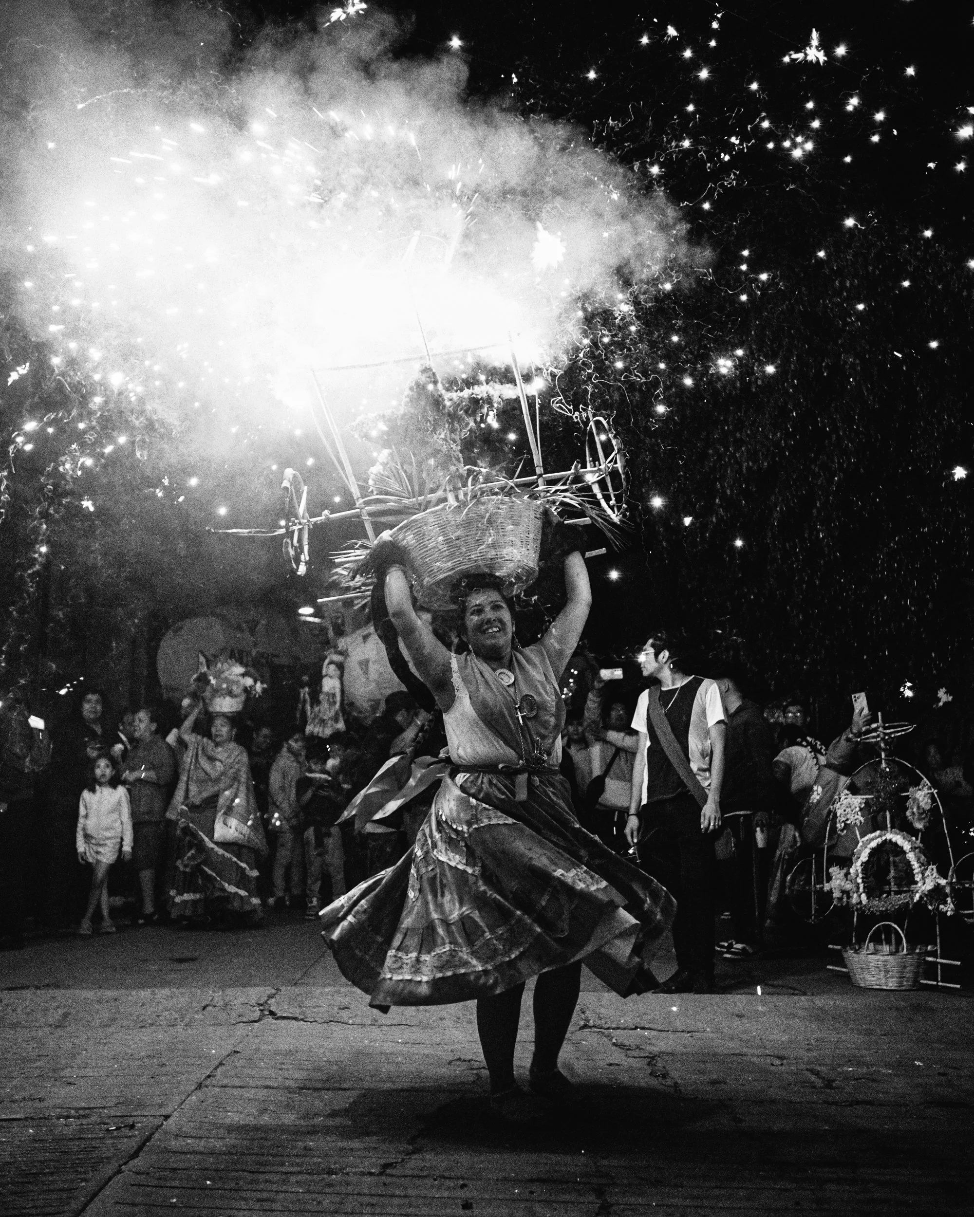 A china oaxaqueña dances at night during a celebration, holding a basket overhead with fireworks in the air around her, with a crowd of people watching.