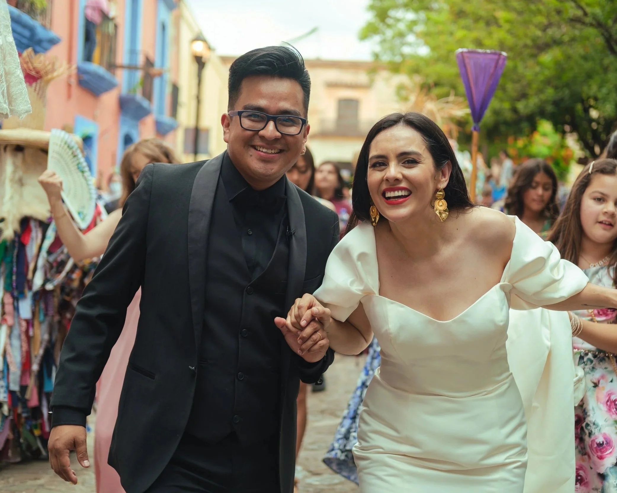 Newlyweds smiling, at a calenda in oaxaca with colorful decorations and guests in the background.