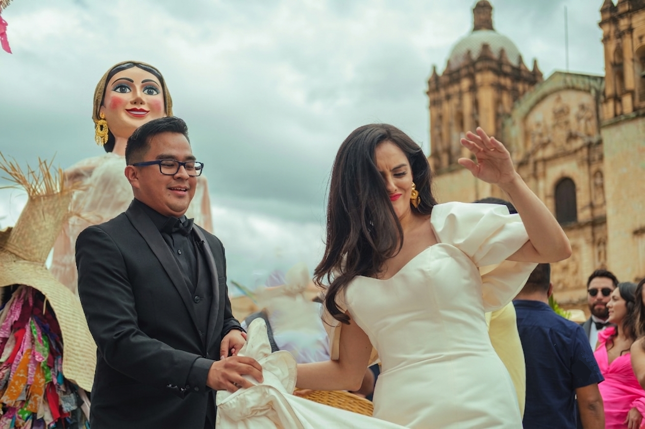 A bride smiling and holding hands with her groom, with traditional dancers wearing colorful costumes and a large puppet figure in the background, Santo Domingo church and historical Oaxacan buildings in the backdrop.