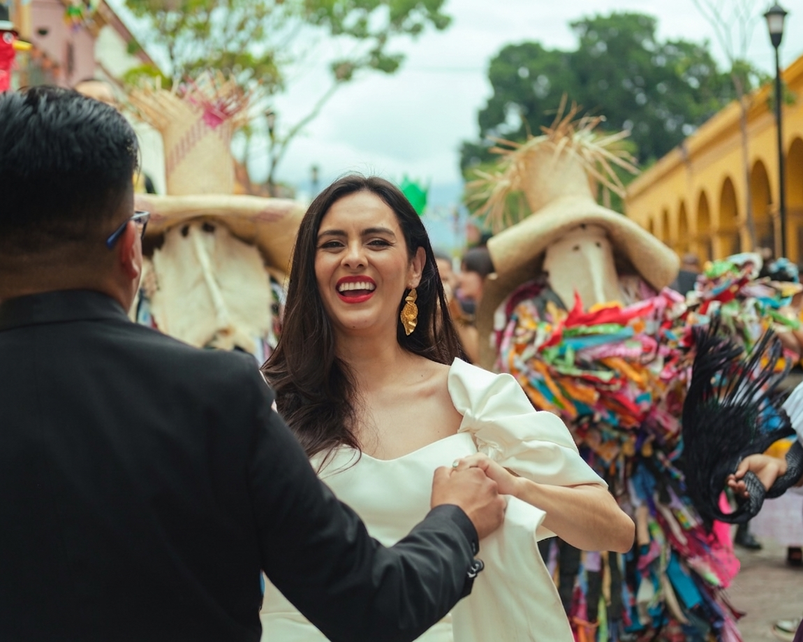 A bride smiling and holding hands with her groom, with traditional dancers wearing colorful costumes and straw hats in the background in a tradicional Oaxacan calenda.
