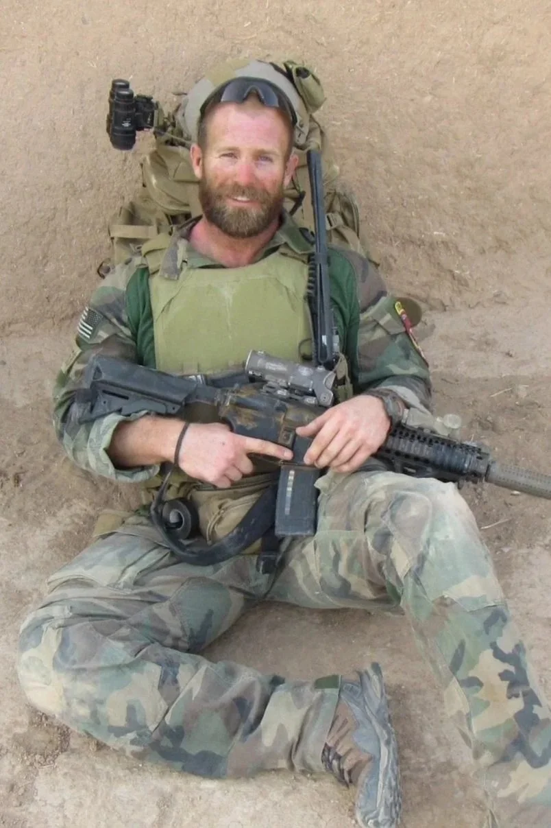A smiling male soldier in military uniform, with camouflage pattern, sitting on the ground against a dirt wall, holding a rifle.