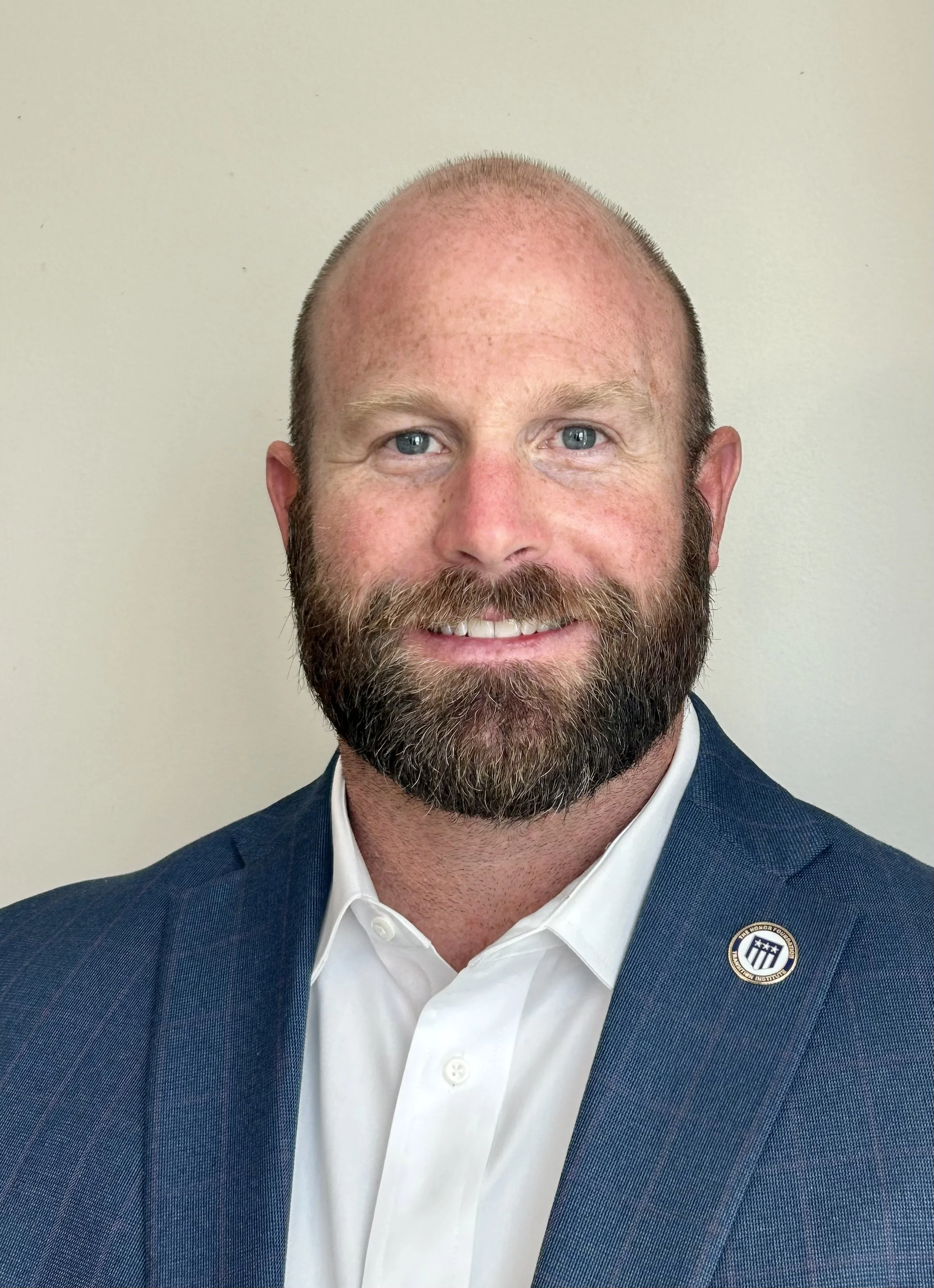 A man with a beard and blue eyes dressed in a navy blazer and white shirt, smiling in front of a plain beige wall, with a round pin badge on his lapel.