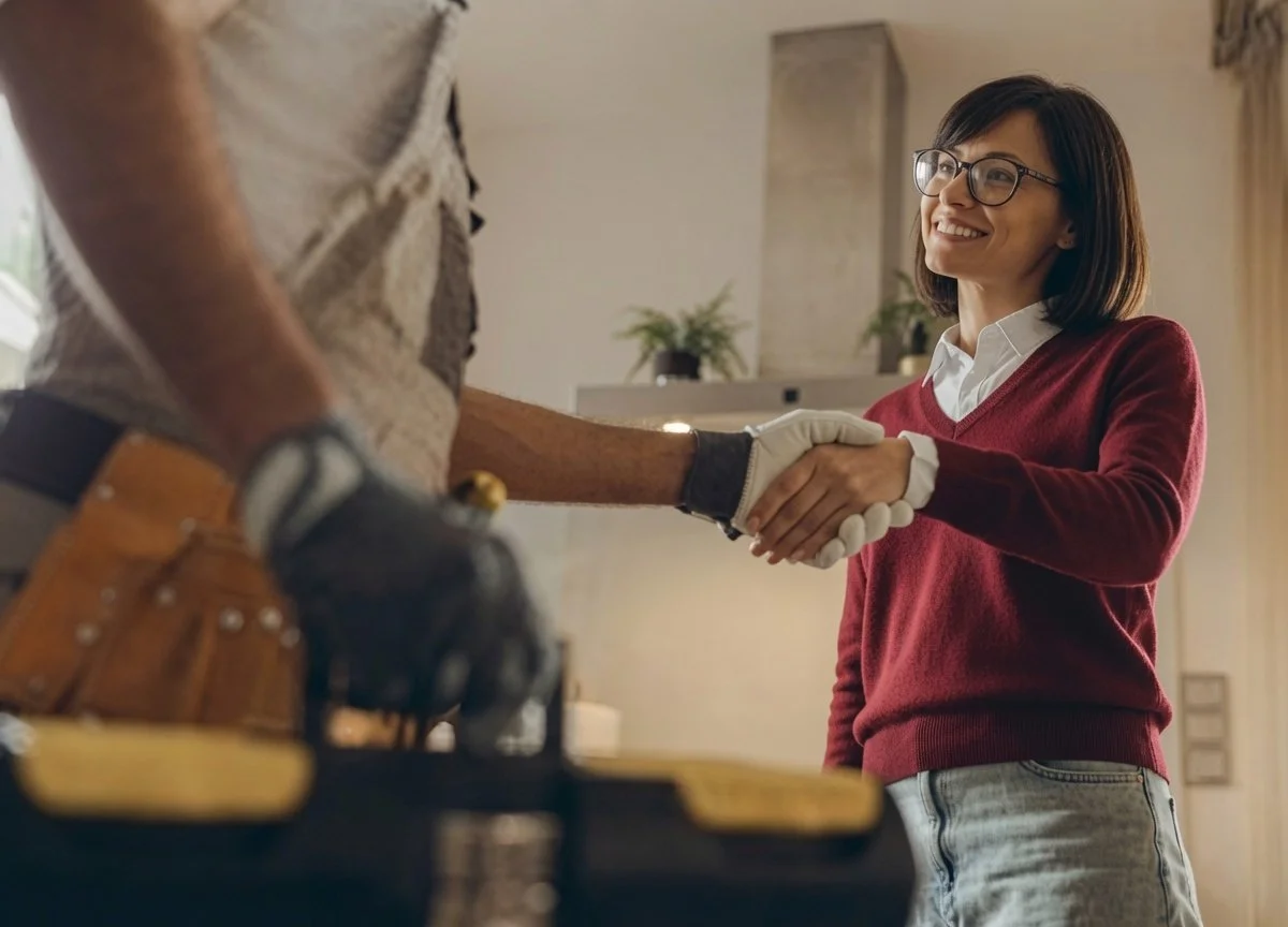 A woman smiling and wearing glasses shakes hands with a man wearing work gloves, in a warmly lit room with a potted plant and a fireplace