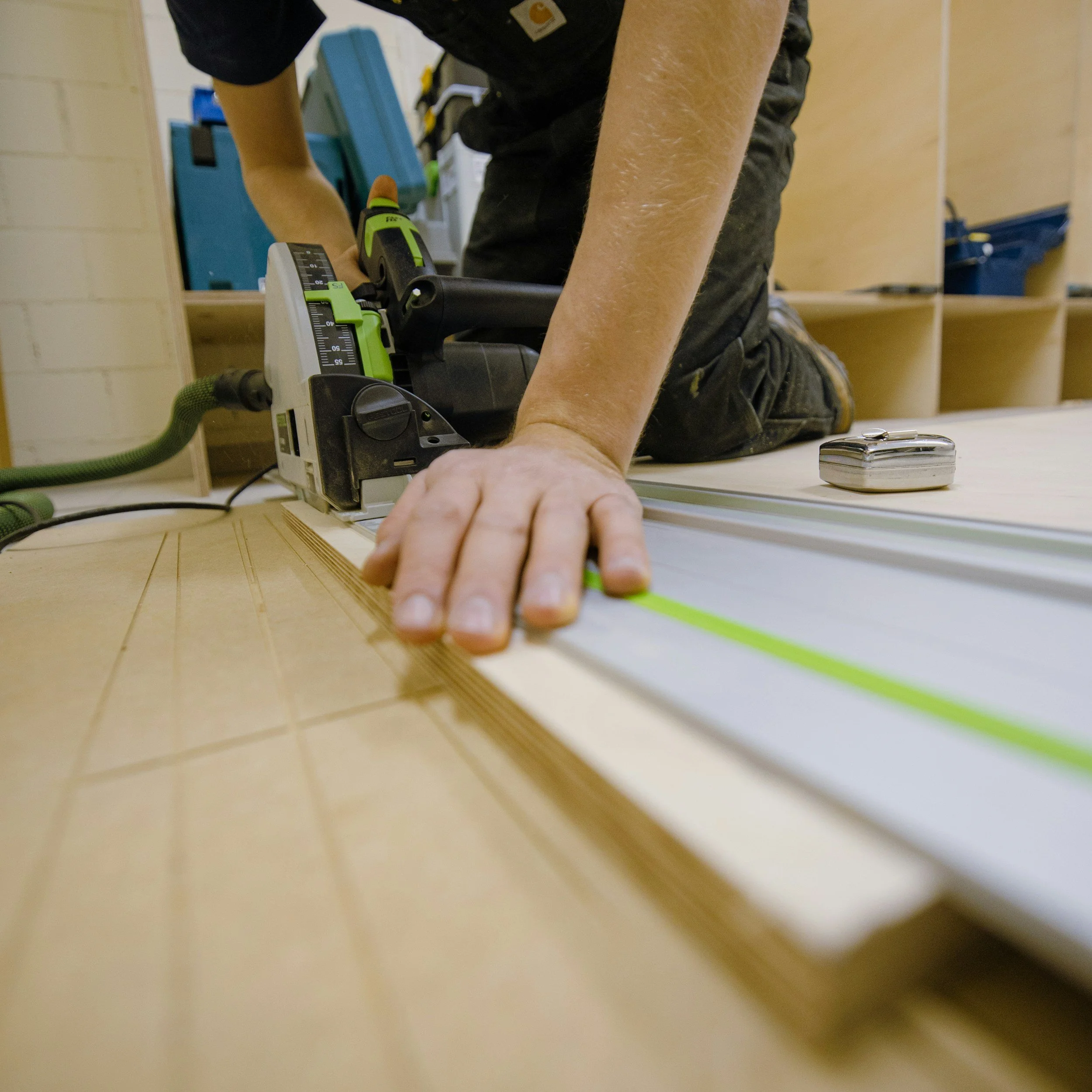 Close-up of a person wearing a black shirt and work gloves, using a green and black power tool on a piece of wood, with woodworking tools and equipment in the background.