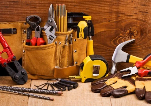 A collection of construction and gardening tools arranged on a wooden surface, including pliers, screwdrivers, a tape measure, hammer, drill bits, and work gloves, with a brown leather tool pouch in the background.