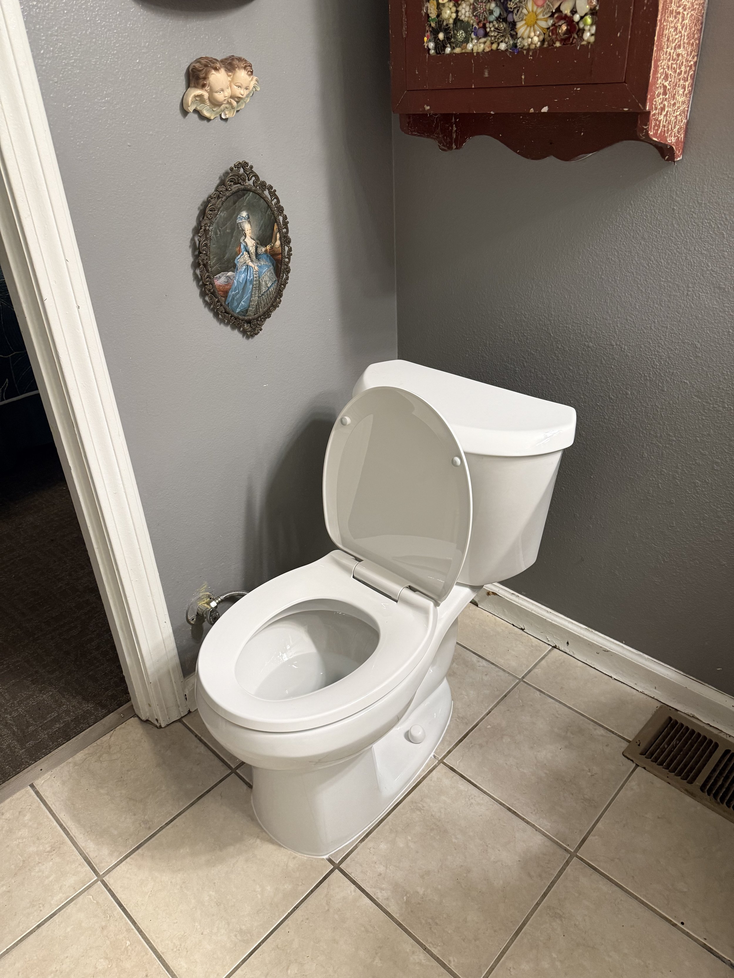 A white toilet with its lid open, placed on a tiled bathroom floor. The wall behind has decorative framed pictures and a wooden cabinet filled with decorative items. There is a floor vent near the toilet.