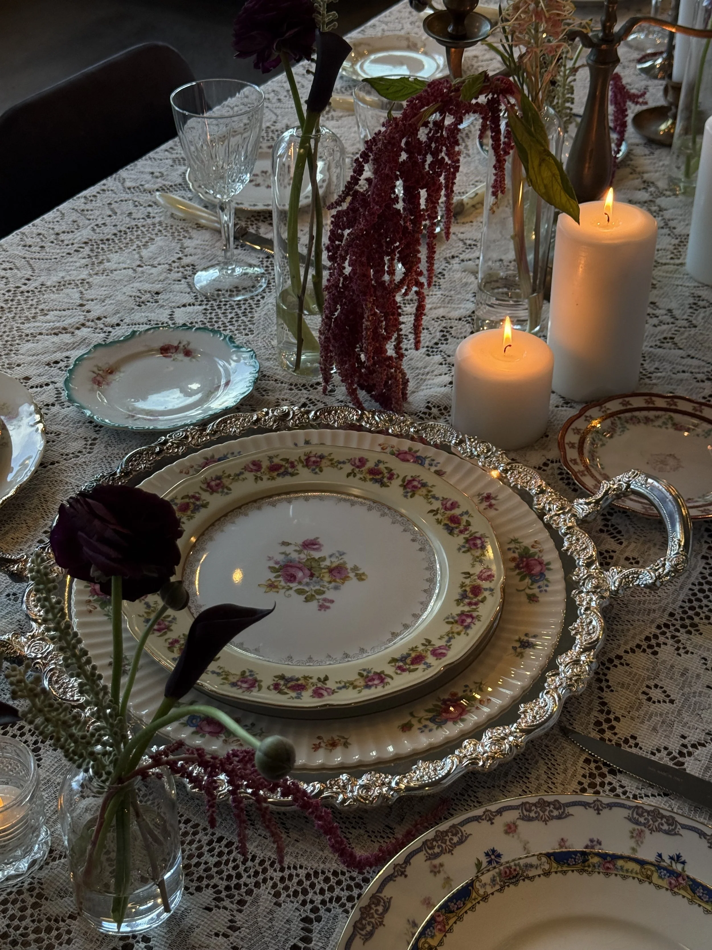 Elegant dining table with floral china plates, crystal glassware, and lit candles, decorated with flowers and a lace tablecloth.