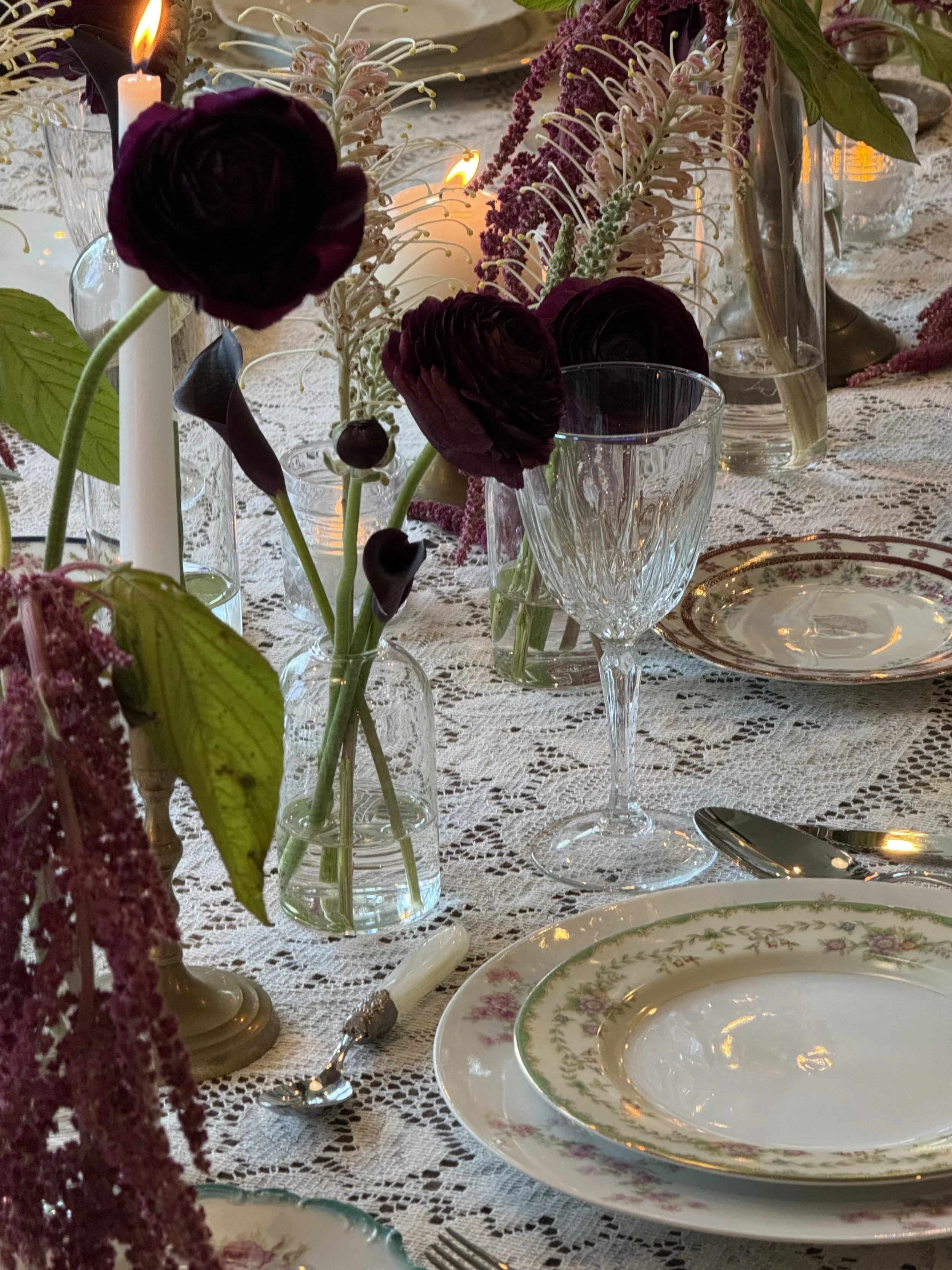 A table decorated with a lace tablecloth, floral arrangement in glass vases, candles, vintage china plates with floral designs, and silverware, creating an elegant setting.